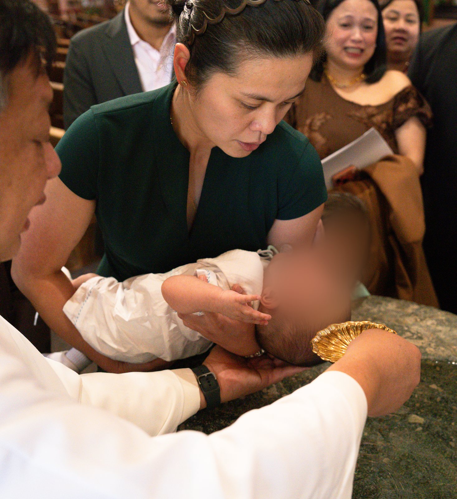 At a stone font, a woman in a green dress holds the baby in a white outfit with the baby's head tipped back while the priest pours water from a gold shell and family look on behind her.
