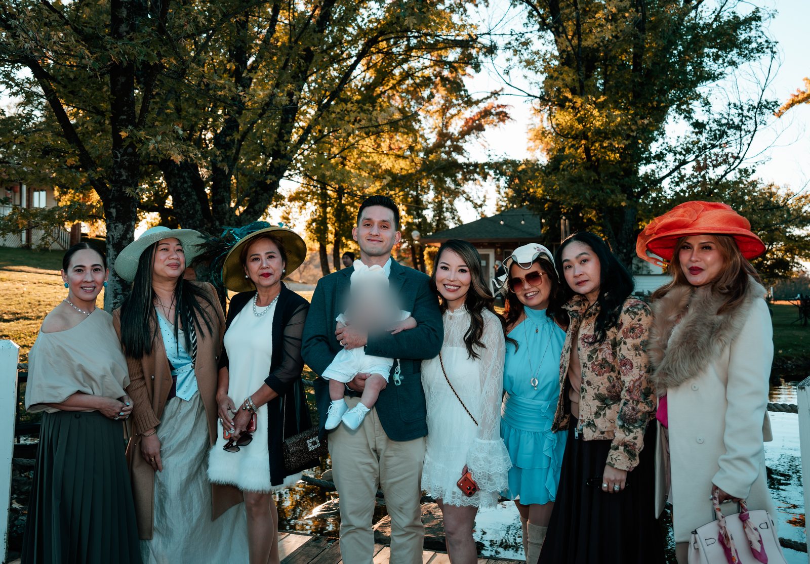 The father holds the baby in a white knit hat among seven women outdoors under autumn trees, several of them wearing wide-brimmed hats, one bright orange.