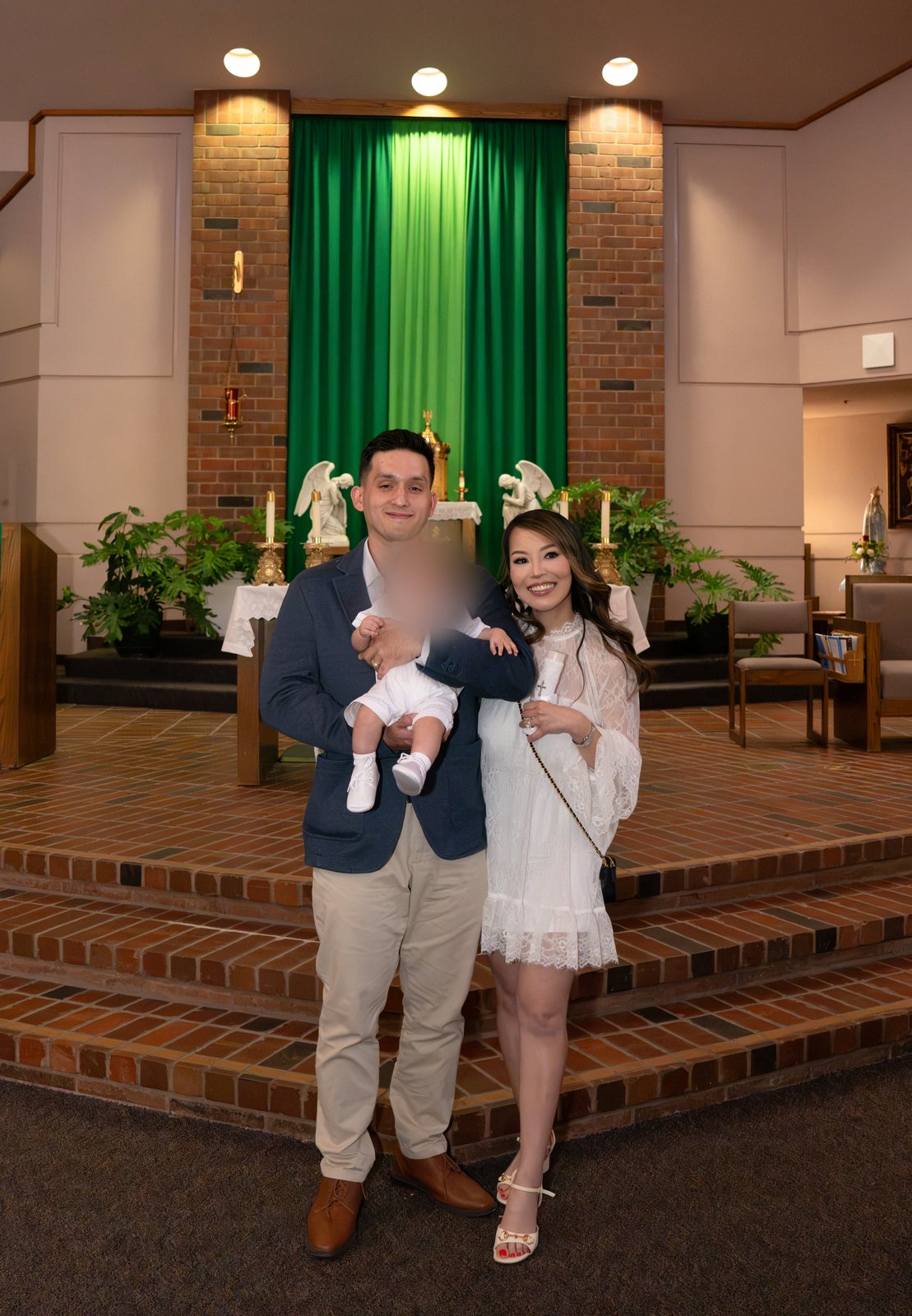 The father holds the baby and the mother holds a white candle as they stand on the brick steps before the altar, between two white angel statues and a green curtain.