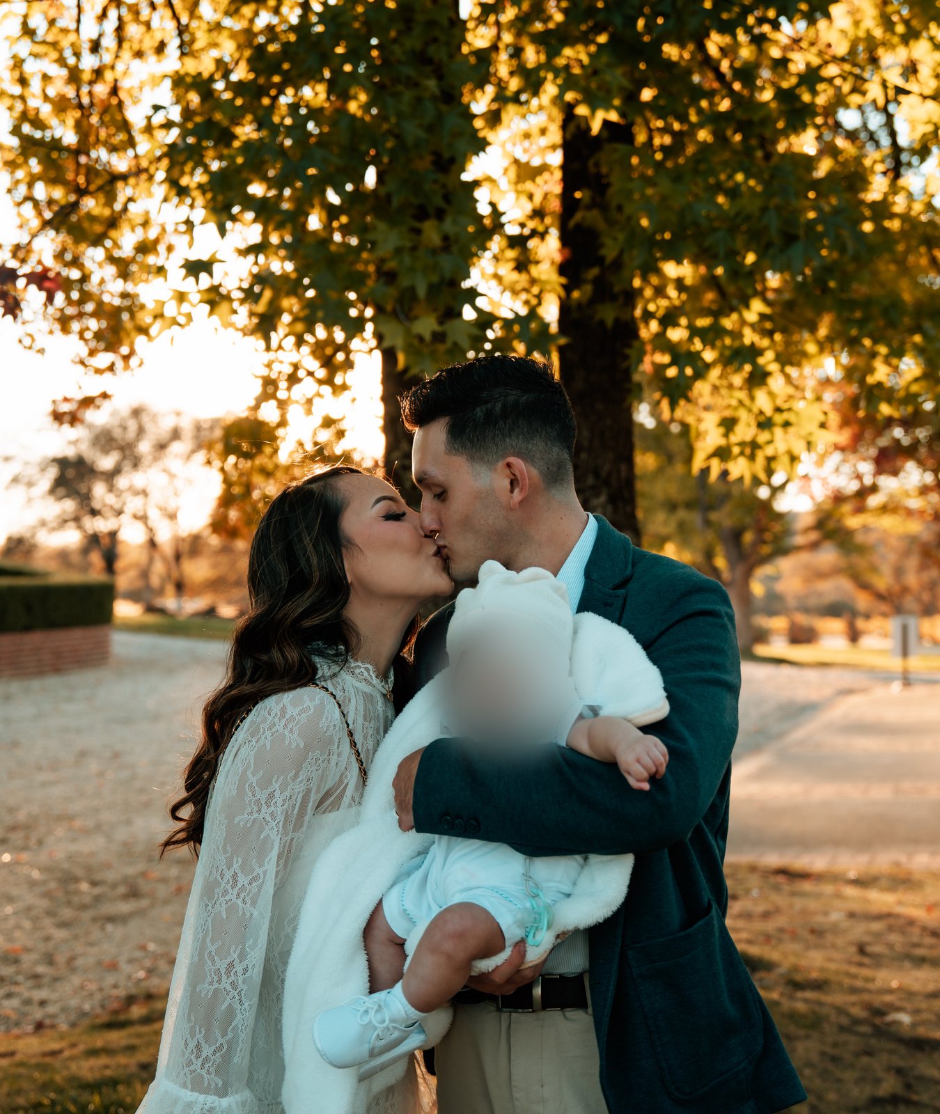 The parents kiss under a tree lit by the low sun while the father holds the baby, wrapped in a white fur blanket and knit hat, against his chest.