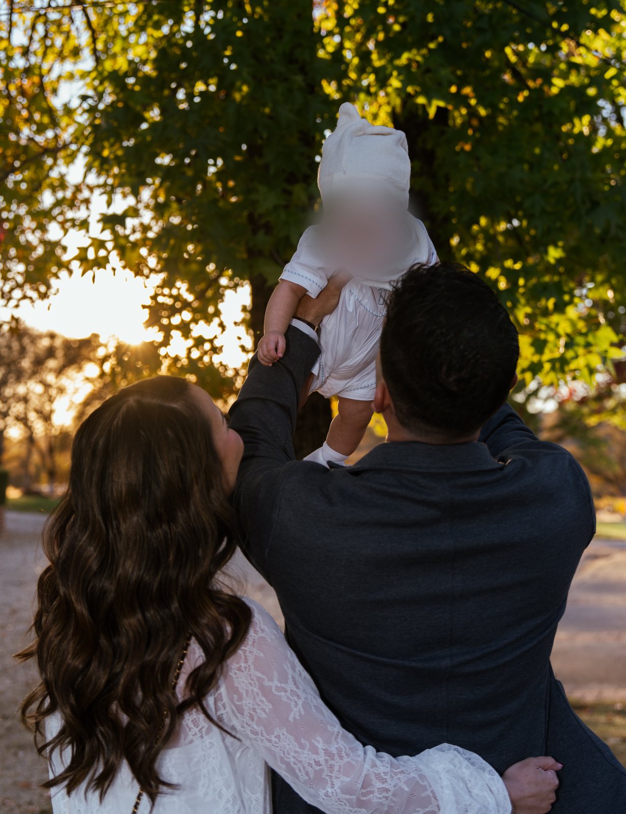 Seen from behind, the father lifts the baby up toward the branches of a sunlit tree while the mother wraps an arm around his waist.