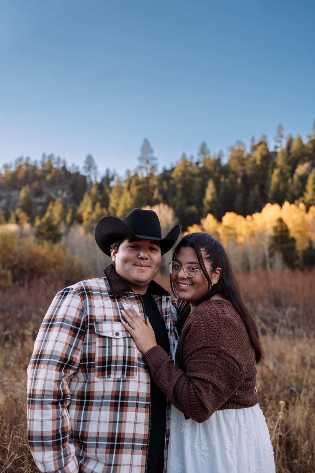 The couple stand close in a dry grass meadow and smile at the camera, her hand on his plaid jacket and his black cowboy hat on, with yellow aspens and pines behind them under a blue sky.