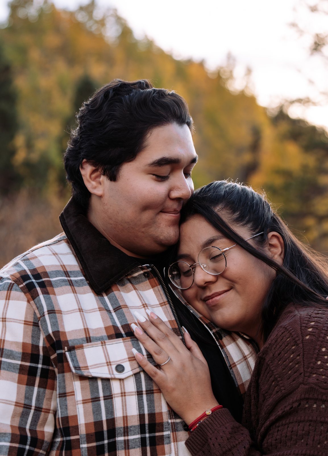 She leans her head against him with her eyes closed and a small smile while he rests his face on her hair, her hand with the ring on his plaid jacket.