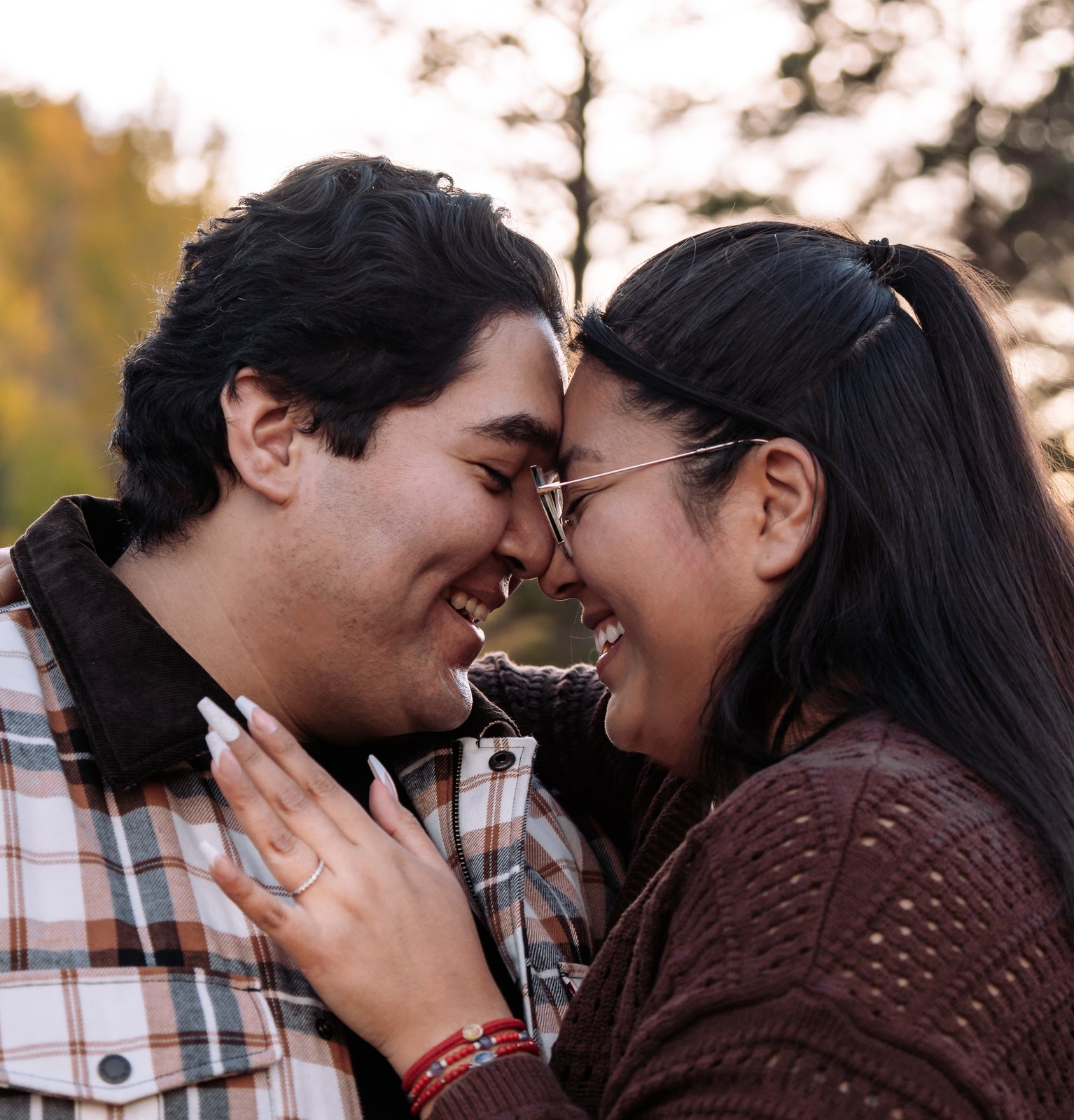 The couple press their foreheads together and laugh with their eyes shut, her ringed hand on his chest and red beaded bracelets on her wrist, with backlit trees behind them.
