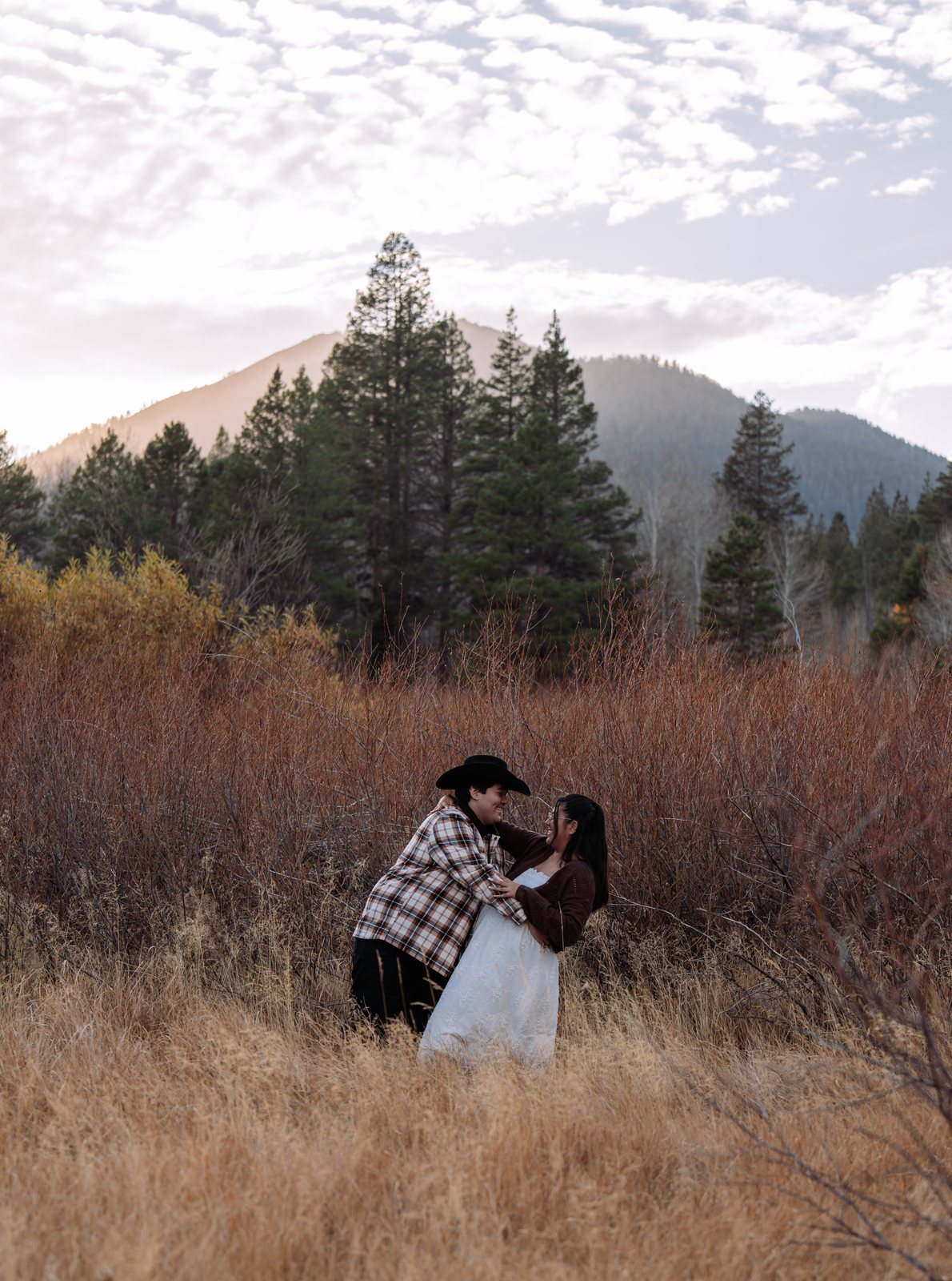 He dips her back in the tall grass and they smile at each other, with red-brown brush, tall pines and a mountain under a sky of small clouds behind them.