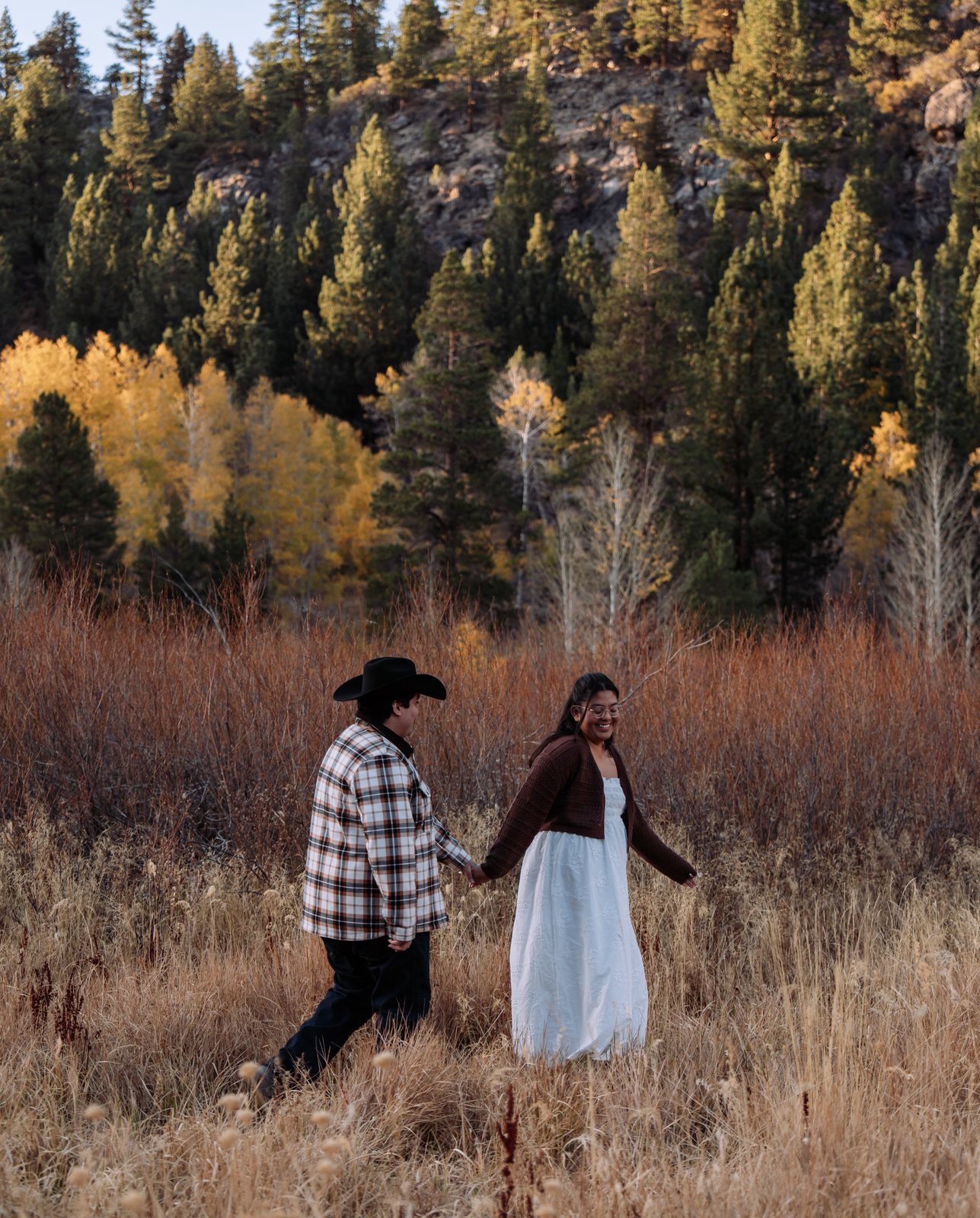 He leads her by the hand through tall dry grass in front of red-brown brush, yellow aspens and a rocky slope of pines, his cowboy hat on and her white dress brushing the grass.