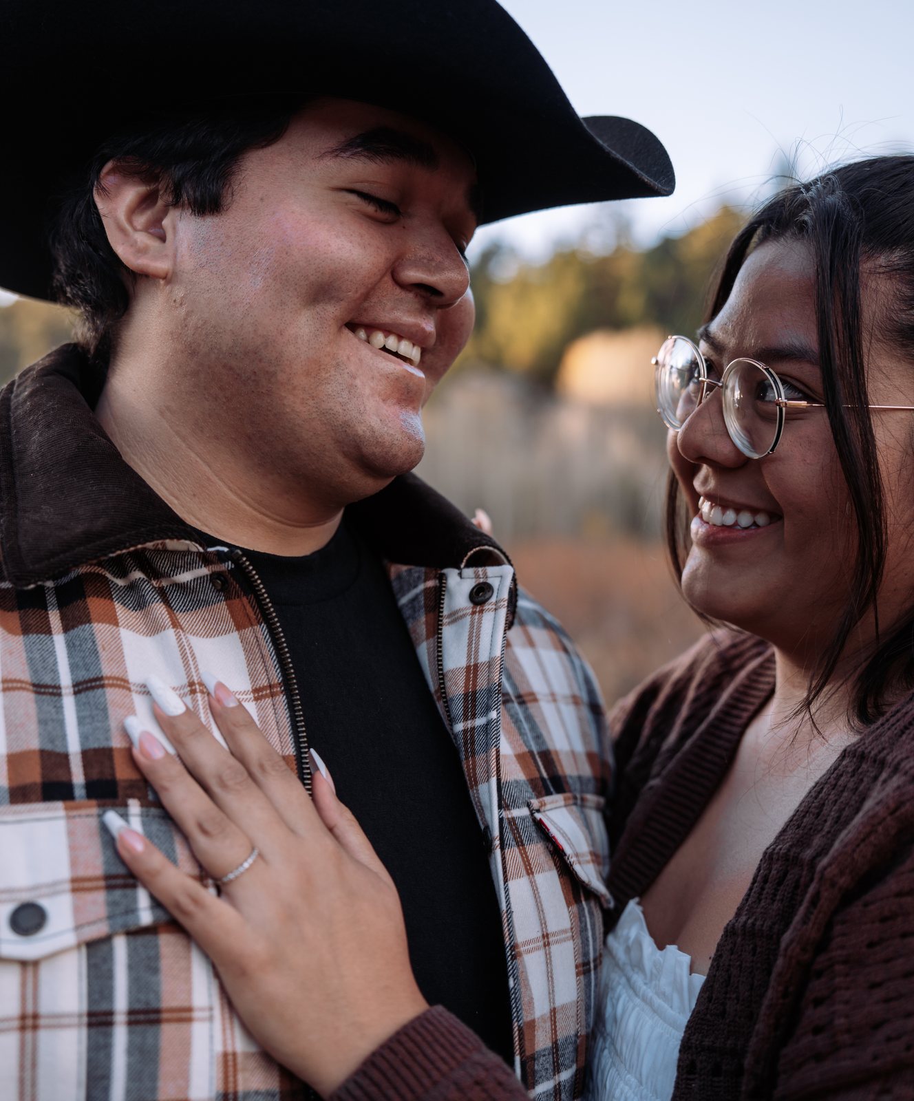 He grins down at her from under his cowboy hat while she smiles up at him through round glasses, her hand with its engagement ring resting on his plaid jacket.