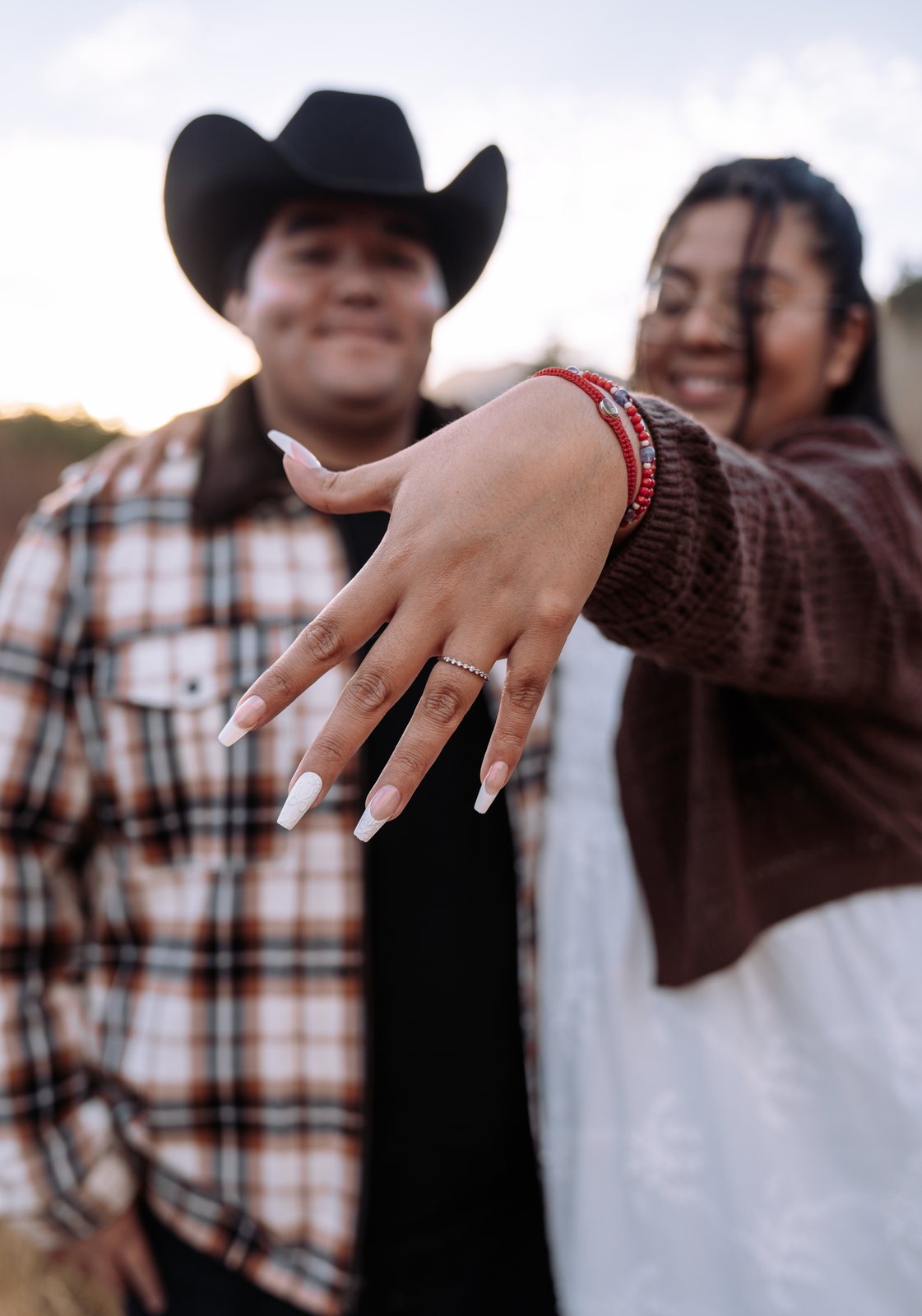She holds her hand out to the camera with the engagement ring in sharp focus while he stands beside her in his cowboy hat, both of them soft and smiling behind it.