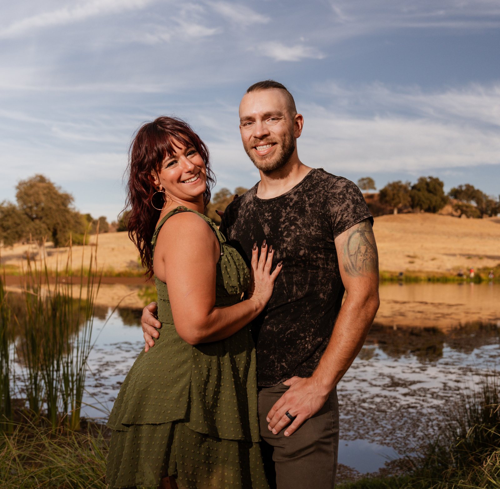 The mother and father stand close at the edge of the pond and smile at the camera, with reeds, still water and dry golden hills with oaks behind them.