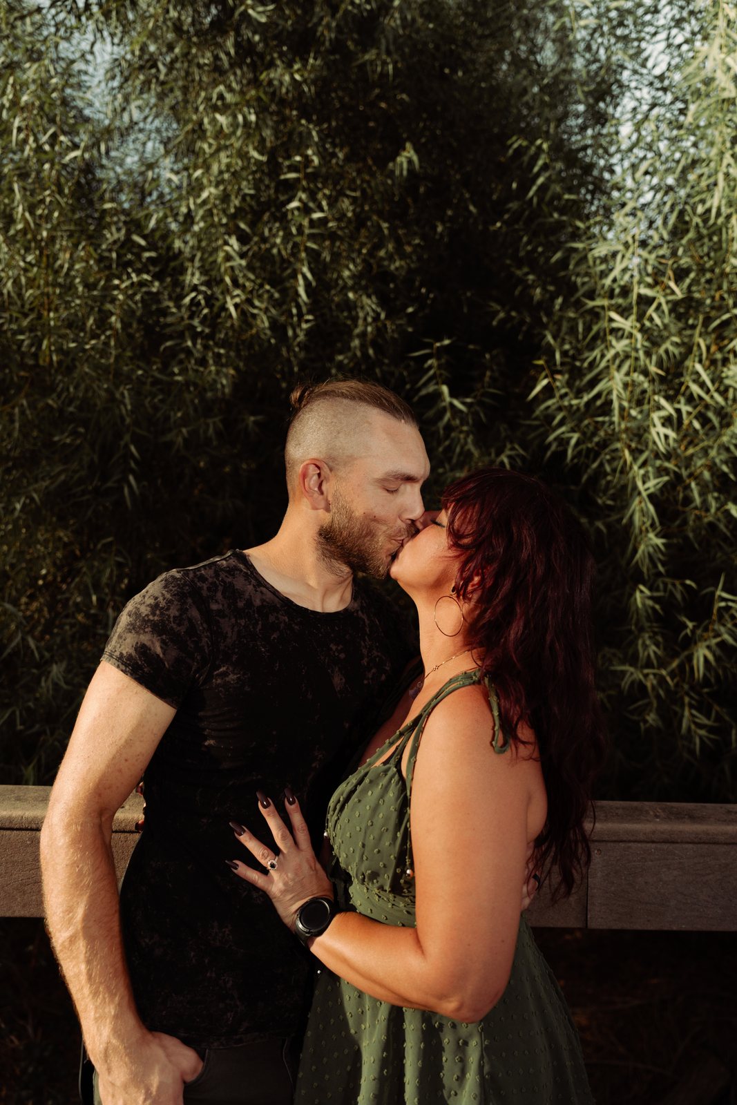 The father and mother kiss with their eyes closed against a wooden railing in front of a dense wall of willow leaves, her hand resting on his chest.