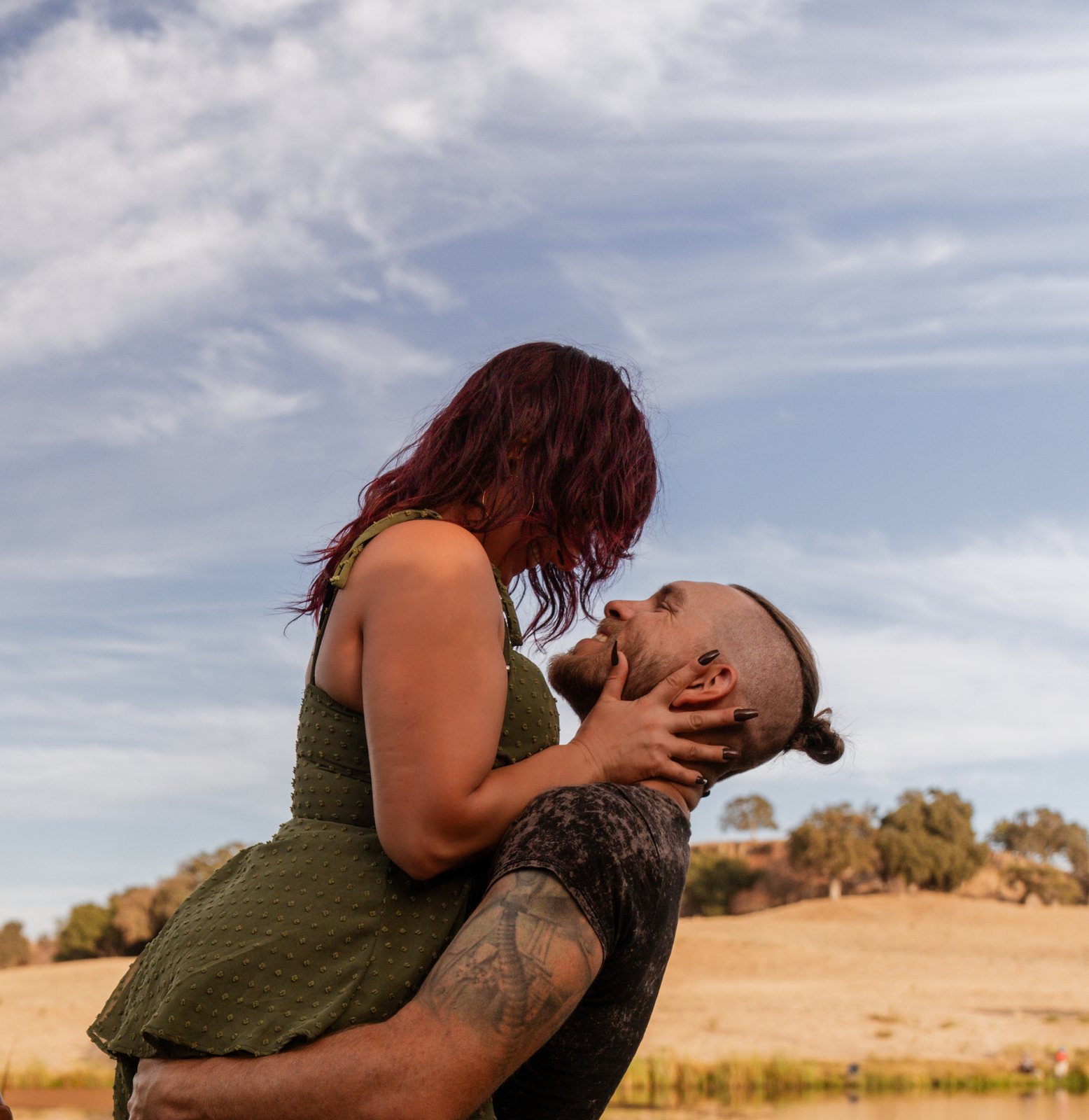 Held up in the father's arms, the mother cups his face in both hands and looks down at him as he smiles up at her against a cloud-streaked sky above the dry hills.