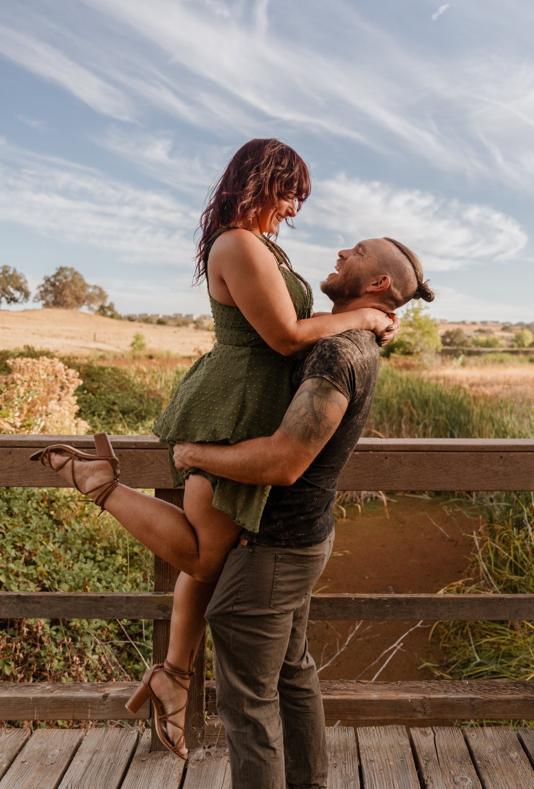 On a wooden bridge, the father lifts the mother off the boards with one heeled sandal kicked up behind her, both laughing as they look at each other under a wide streaky sky.