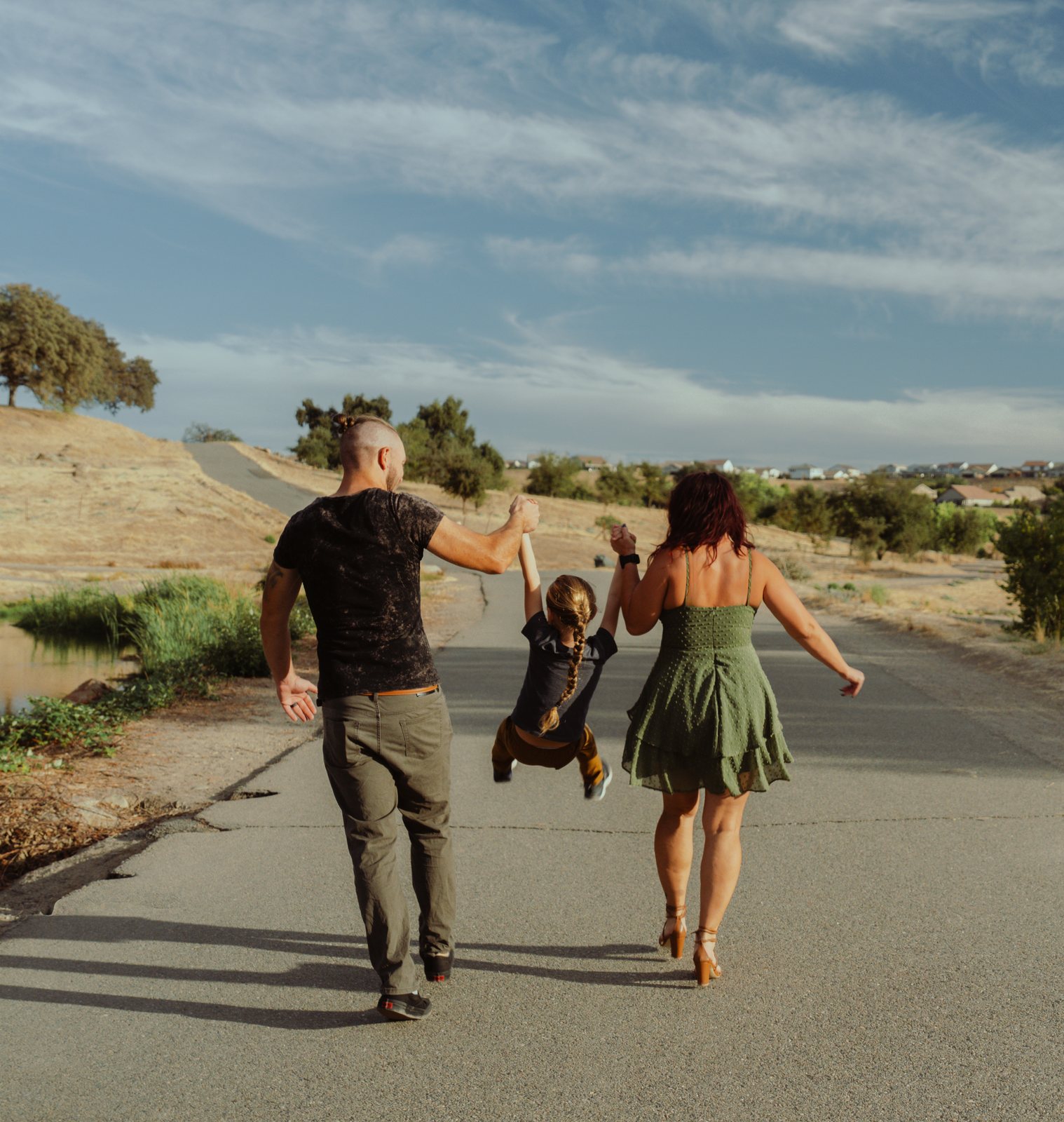 Seen from behind, the parents swing their son between them by the hands, his braid down his back and his feet off the ground, on a paved path through dry hills with a pond at the left.