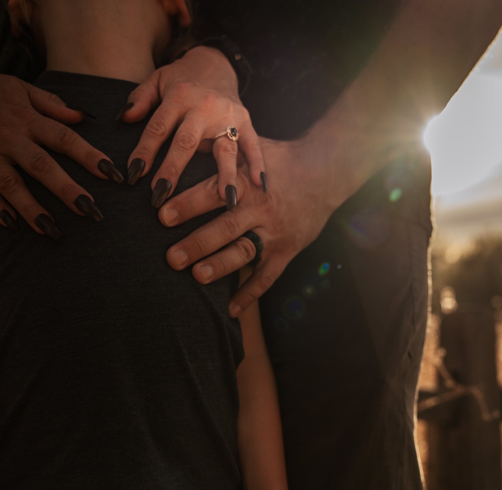 The parents' hands rest on their son's shoulders, the mother's dark nails and ring against his grey T-shirt and the father's dark ring below, with low sun flaring at the edge.