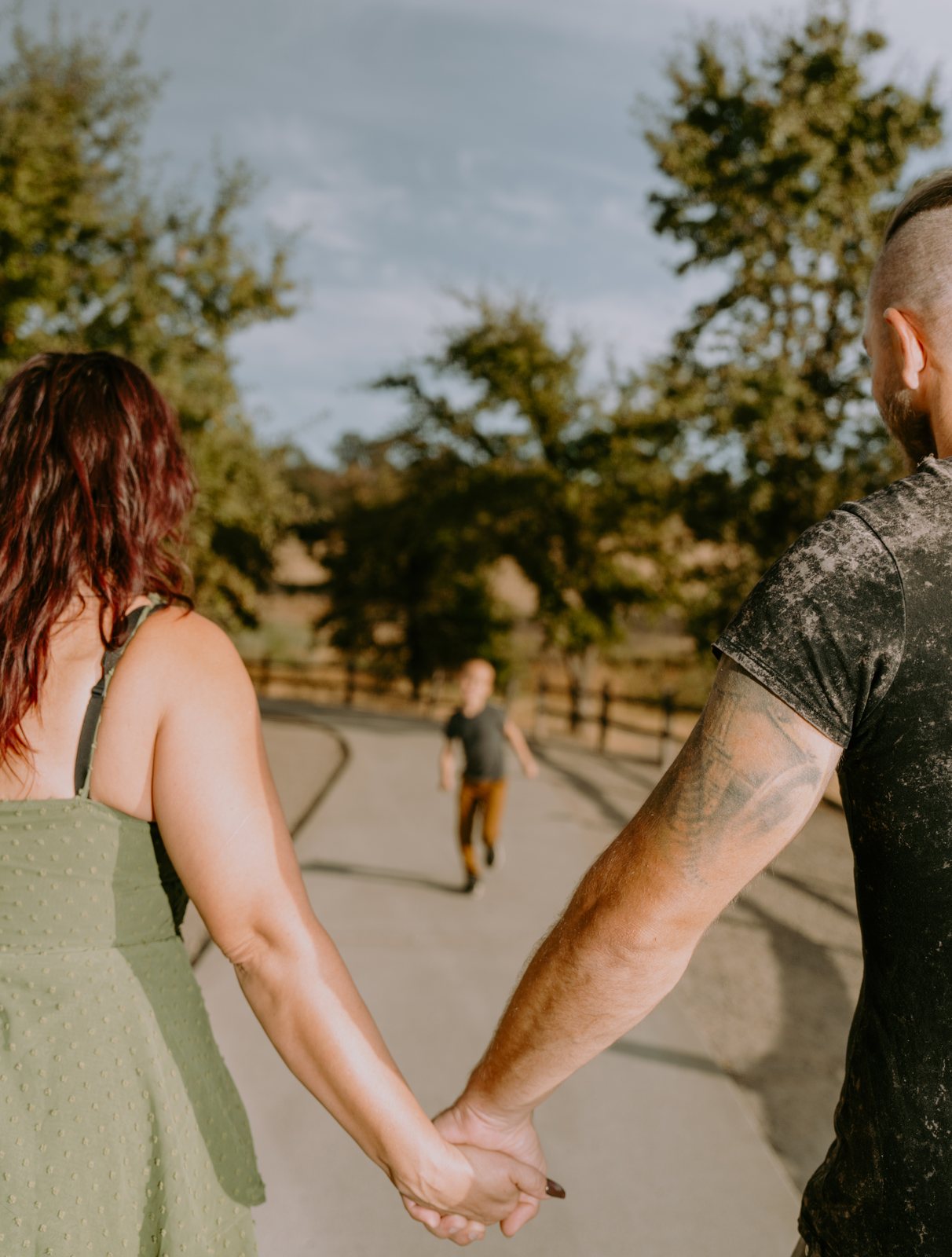 Seen from behind, the mother and father hold hands on a curving paved path while their son, out of focus in a grey T-shirt and brown pants, runs toward them past a wooden fence.