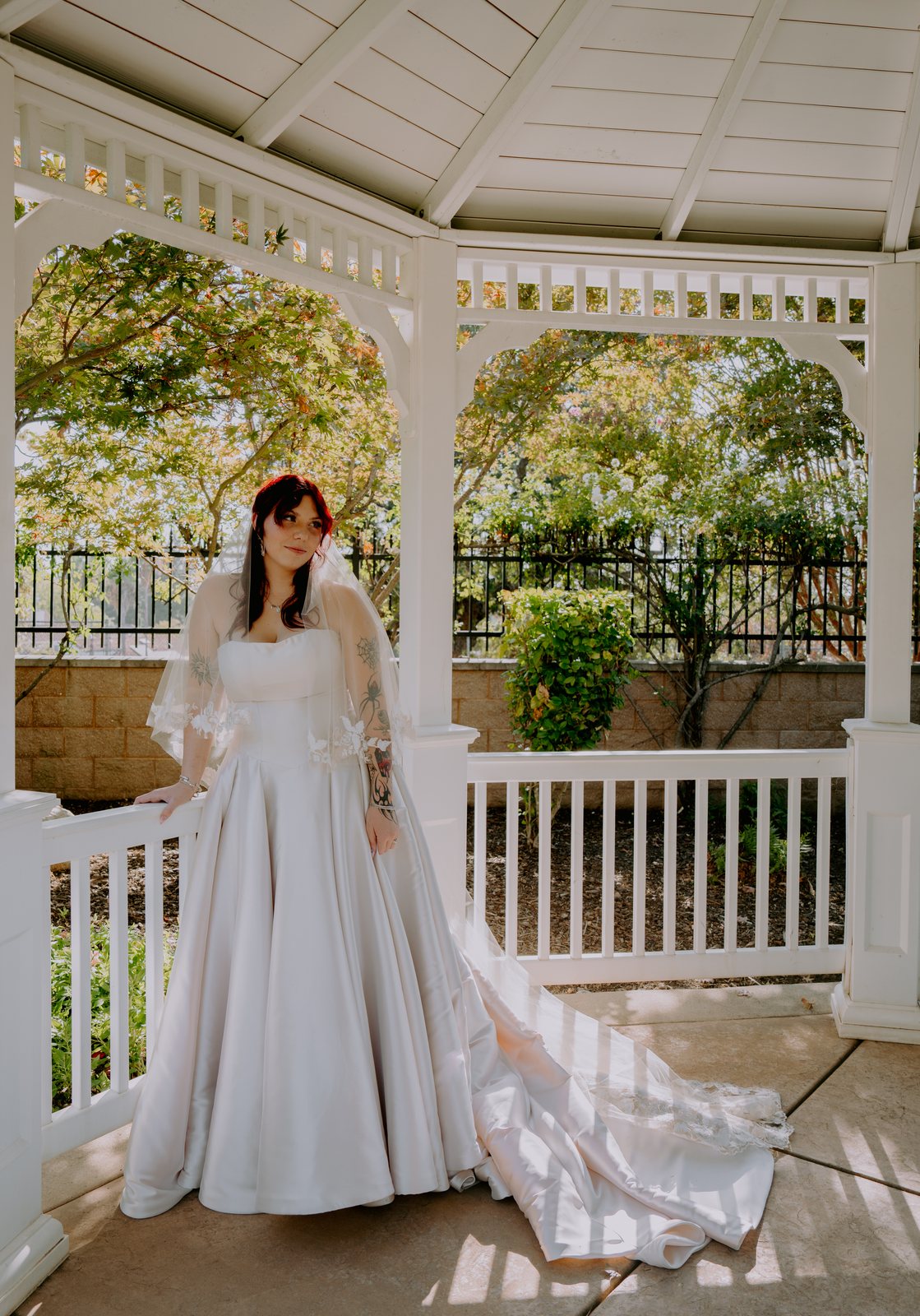 The bride stands alone in the gazebo with one hand on the white railing, her satin train fanned out behind her across stripes of sun on the floor.