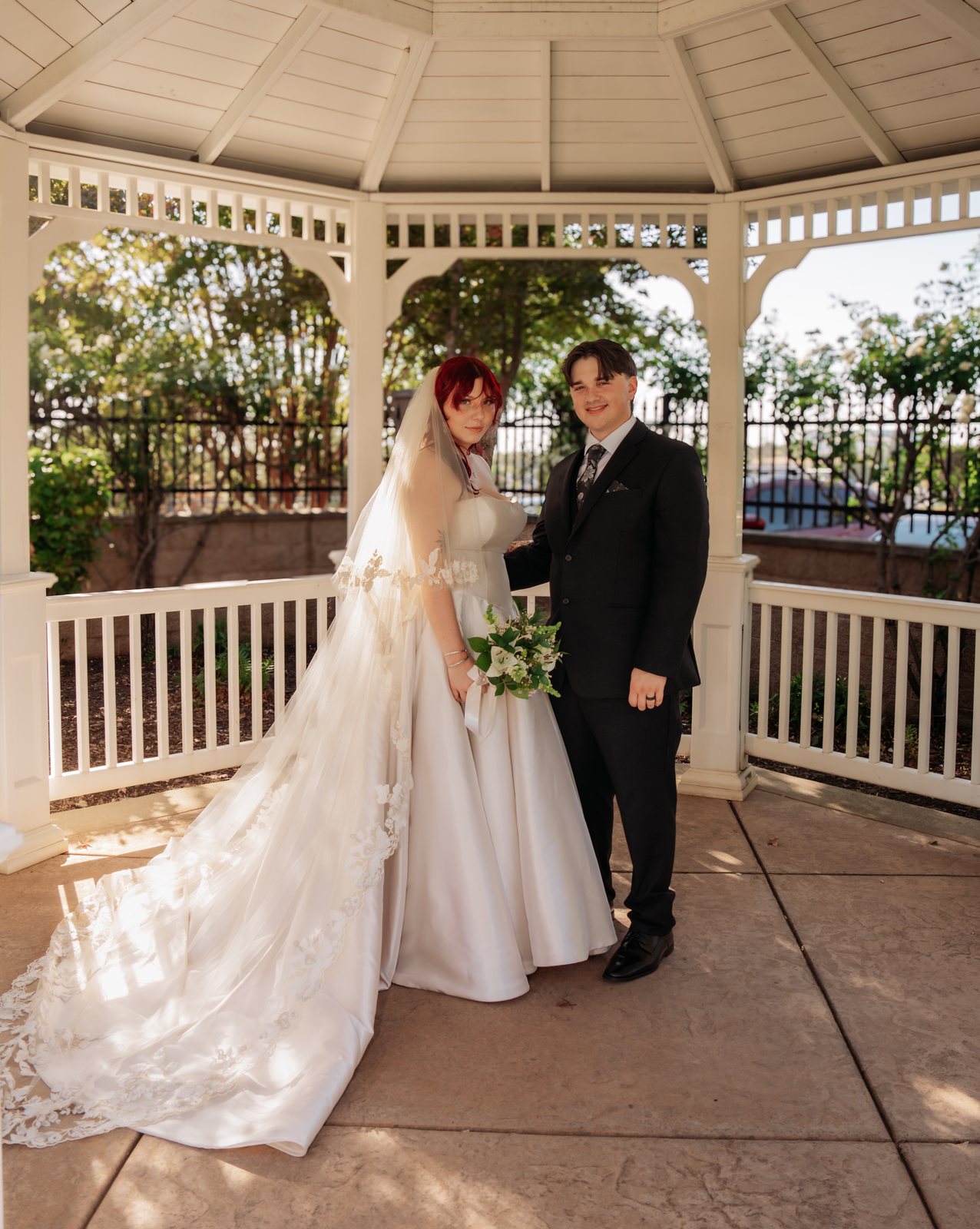 The bride and groom stand side by side in the middle of the gazebo smiling at the camera, her bouquet at her waist and her lace-edged veil and train trailing across the floor.