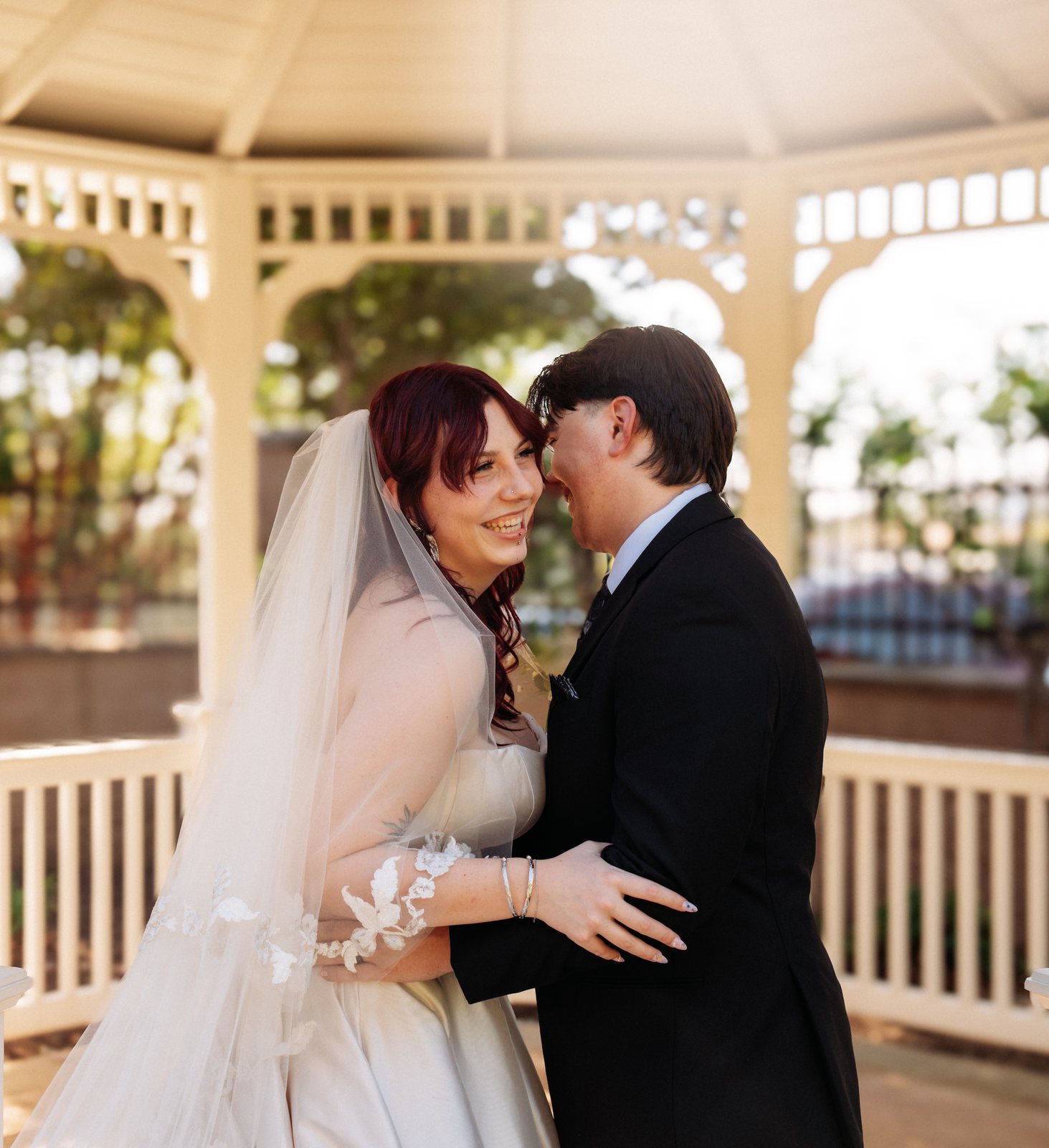 The bride laughs with her cheek against the groom's face as he grins with his eyes closed, her hand on his arm, under the roof of a white gazebo.