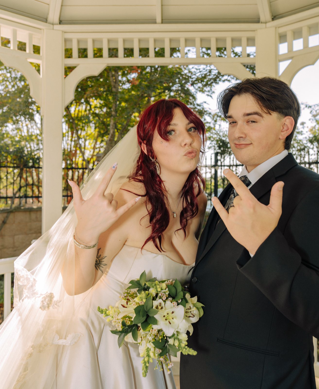The bride puckers her lips at the camera and the groom grins as both hold up a rock-on hand sign, her bouquet of lilies held low in front of her gown.