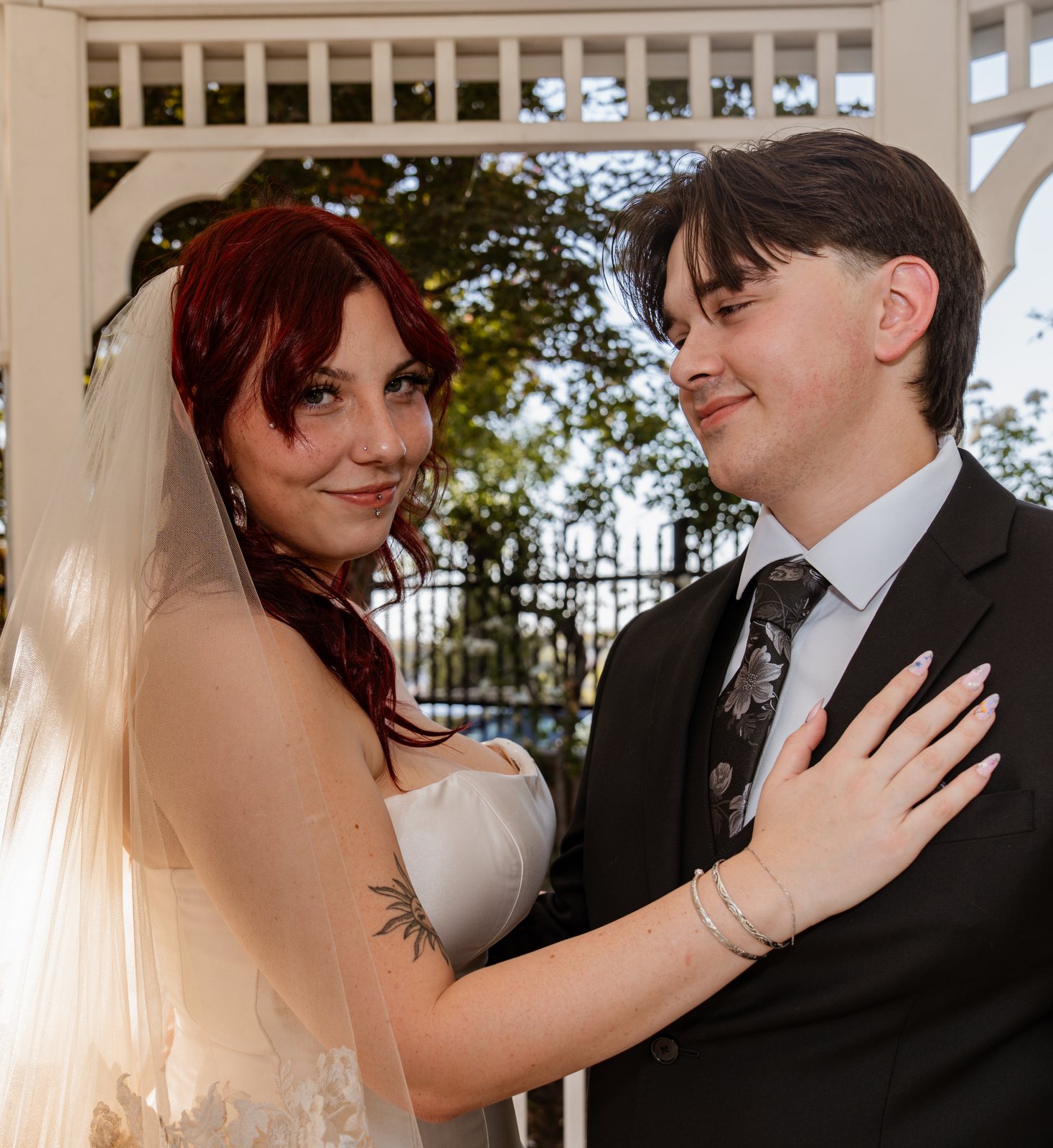 The bride rests her hand on the groom's chest and smiles at the camera while he smiles down at her, in front of the gazebo's white trim and an iron fence.