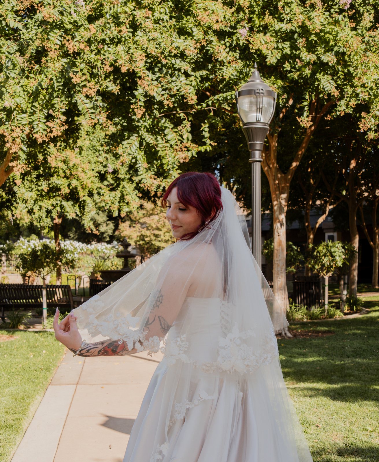 The bride turns on a park path and looks back over her shoulder as she holds out the edge of her veil, under trees beside a tall lamp post.