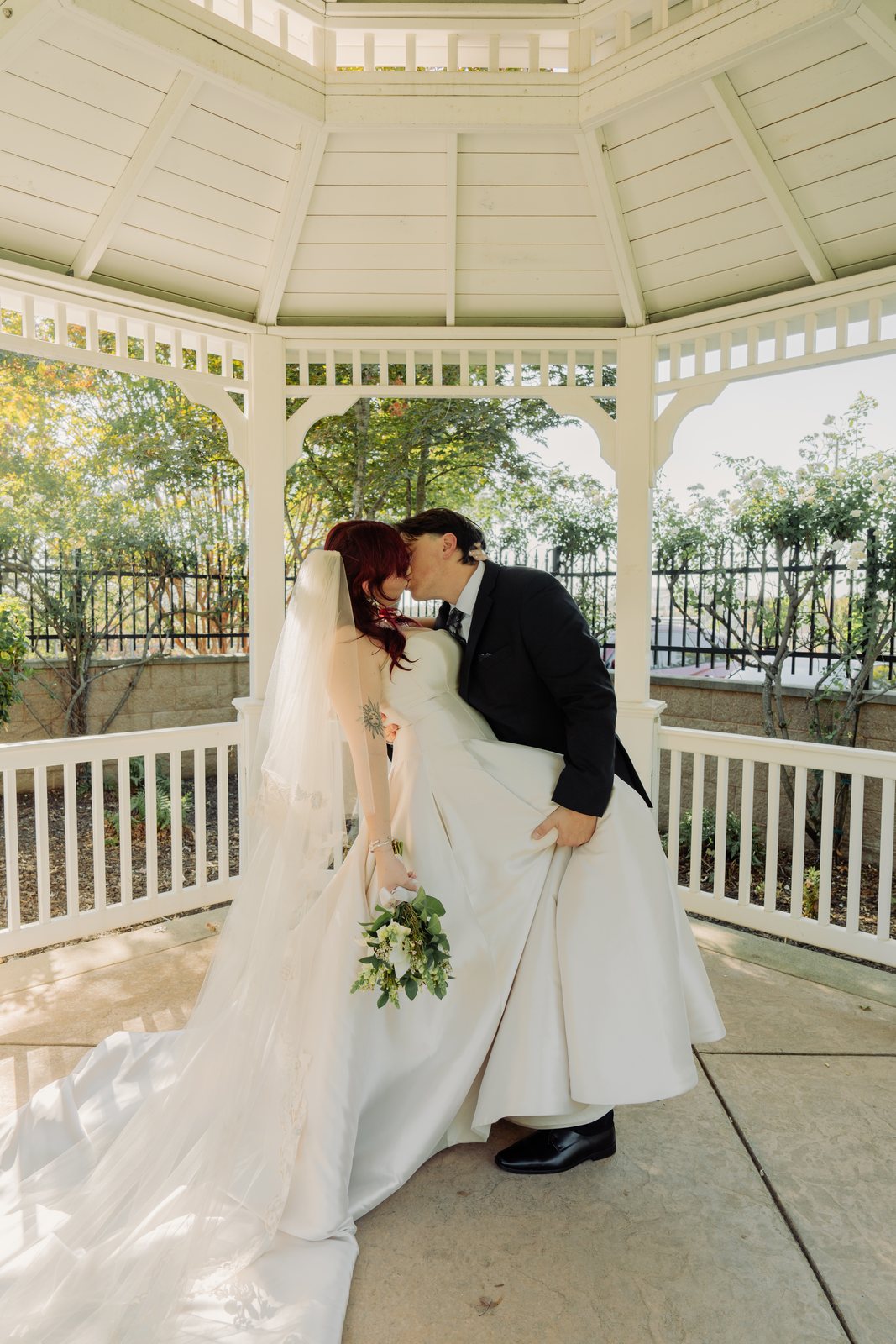 The groom dips the bride and kisses her in the middle of the gazebo, holding out the side of her satin skirt, her bouquet in her hand and her veil trailing across the floor.