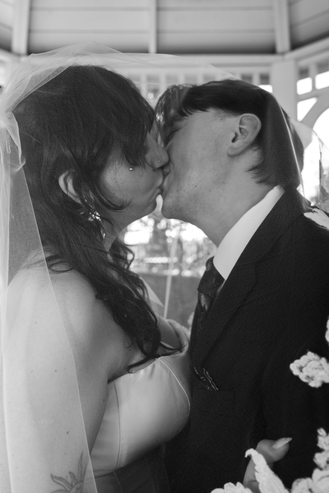 In black and white, the bride and groom kiss with the edge of her veil over them, white lilies from her bouquet at the corner of the frame.