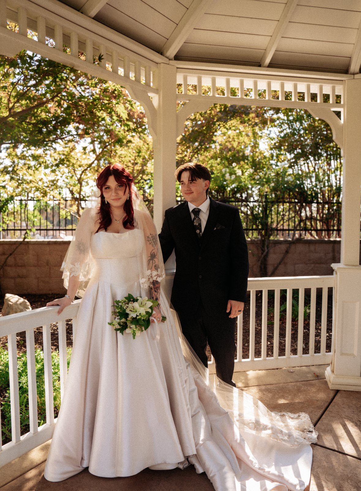 The bride leans on the gazebo railing with a bouquet of white lilies while the groom stands just behind her shoulder, her train spread across the sunlit floor.