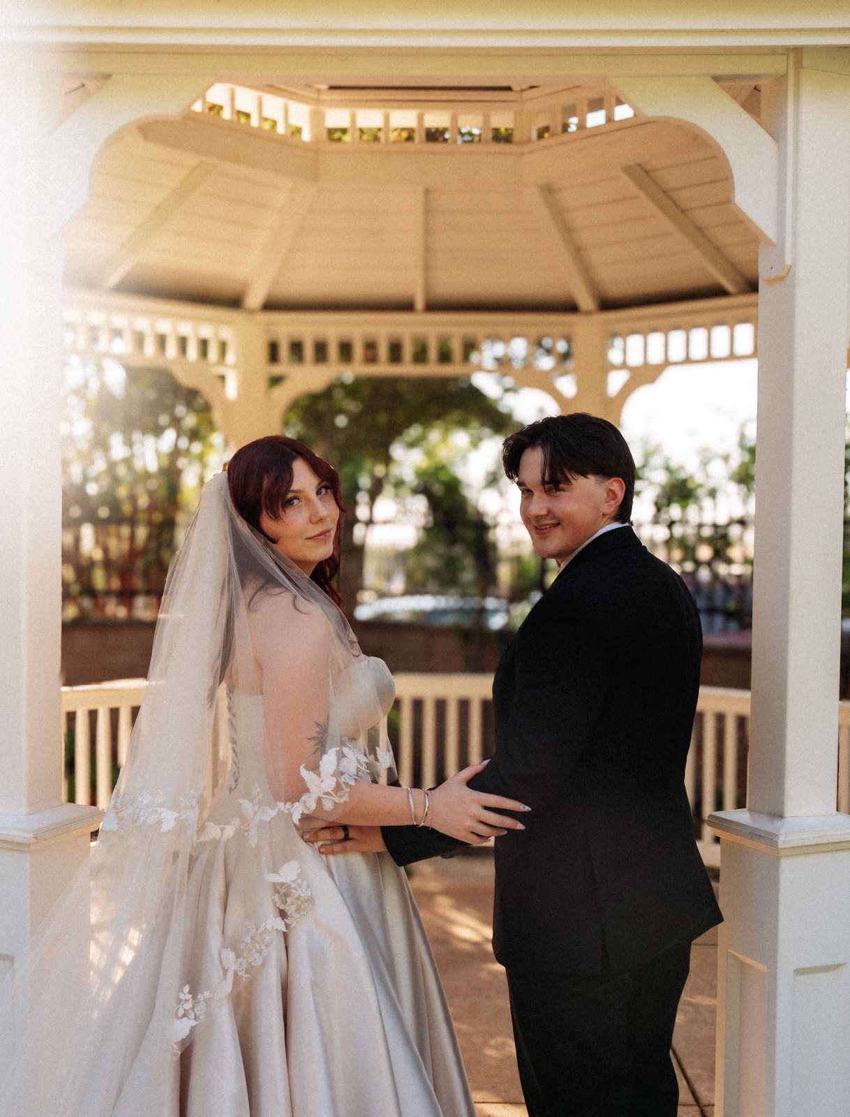 In a white wooden gazebo, the bride and groom stand half turned away and look back over their shoulders at the camera, his hand at her waist and hers on his arm.