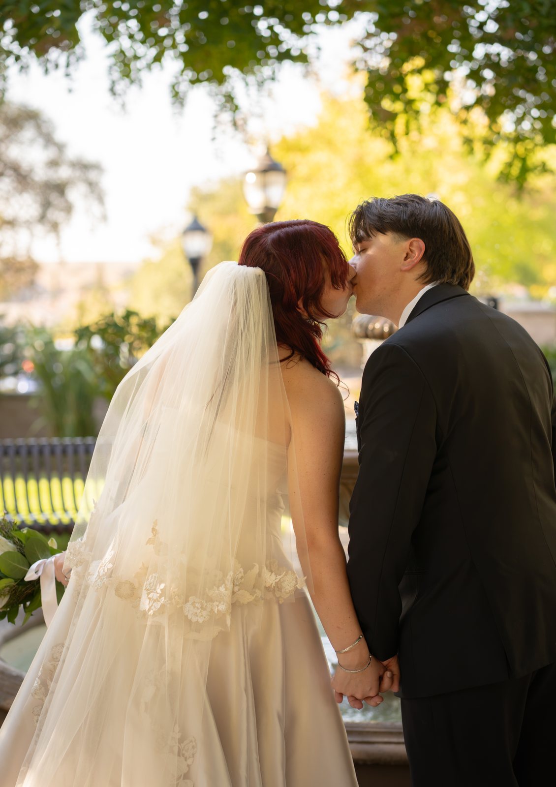 Seen from behind, the bride in a long lace-edged veil holds the groom's hand as he leans in to kiss her at the edge of a stone fountain, with lamp posts and yellowing trees beyond.