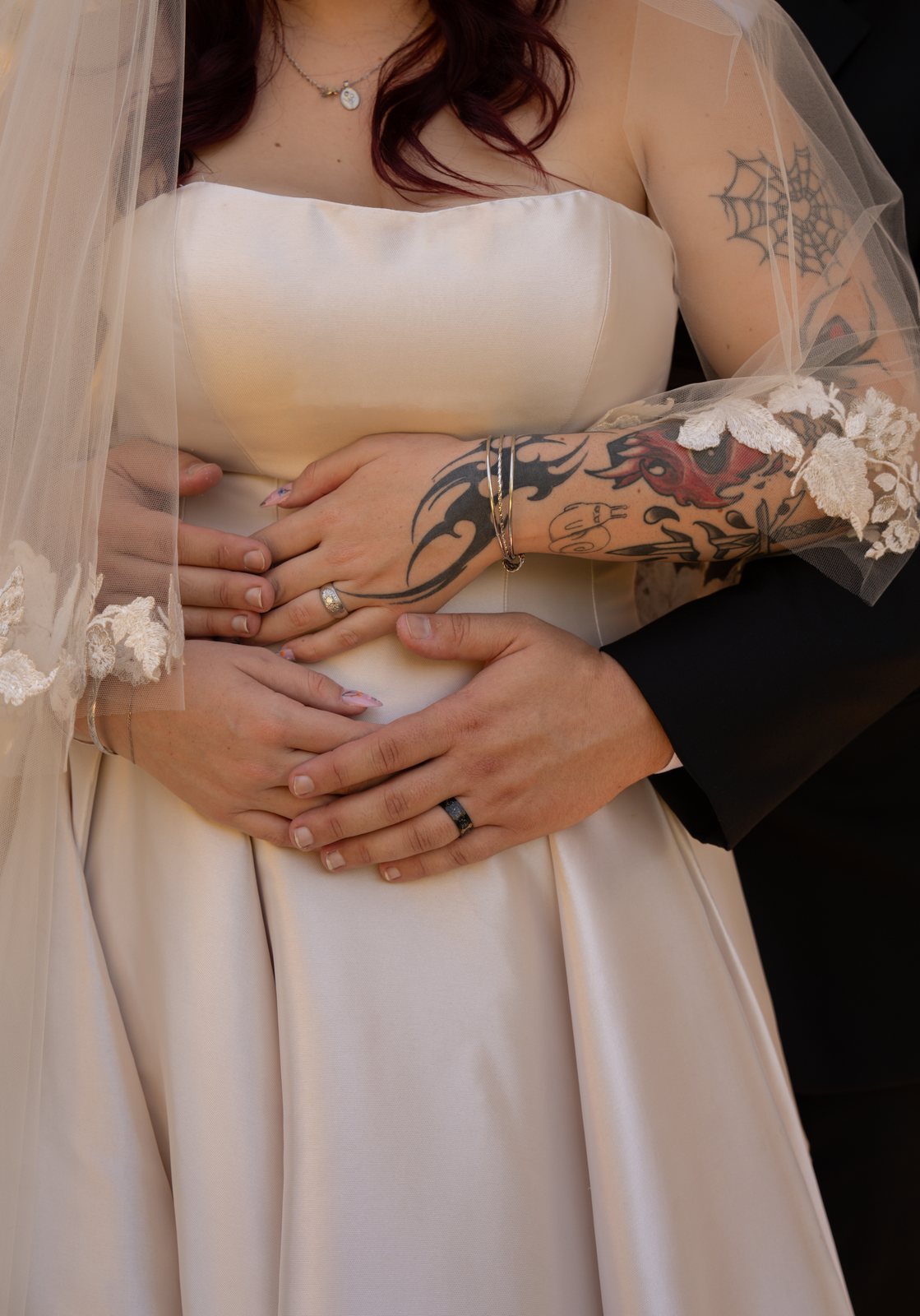 Four hands layered at the bride's waist over her satin dress, her silver ring and the groom's dark band both showing, beside her tattooed arm and lace-edged veil.