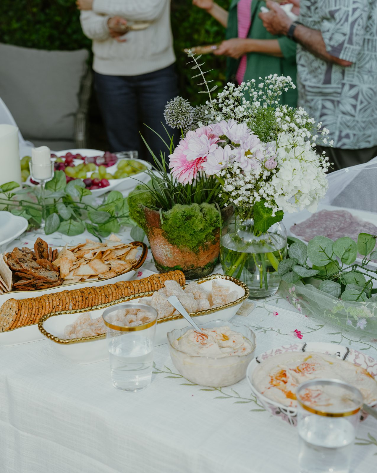 A party table with crackers, pita chips, bowls of dip and glasses of water around a centerpiece of pink gerberas, baby's breath and moss, with guests standing behind it.