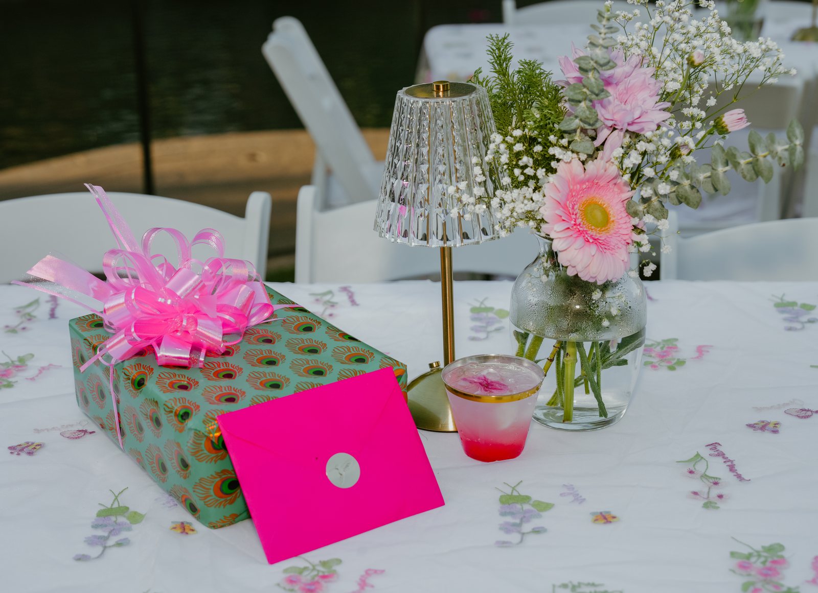 A gift wrapped in peacock-feather paper and tied with a pink bow sits on an embroidered tablecloth beside a pink envelope, a glass lamp, a pink drink and a vase of gerbera daisies.