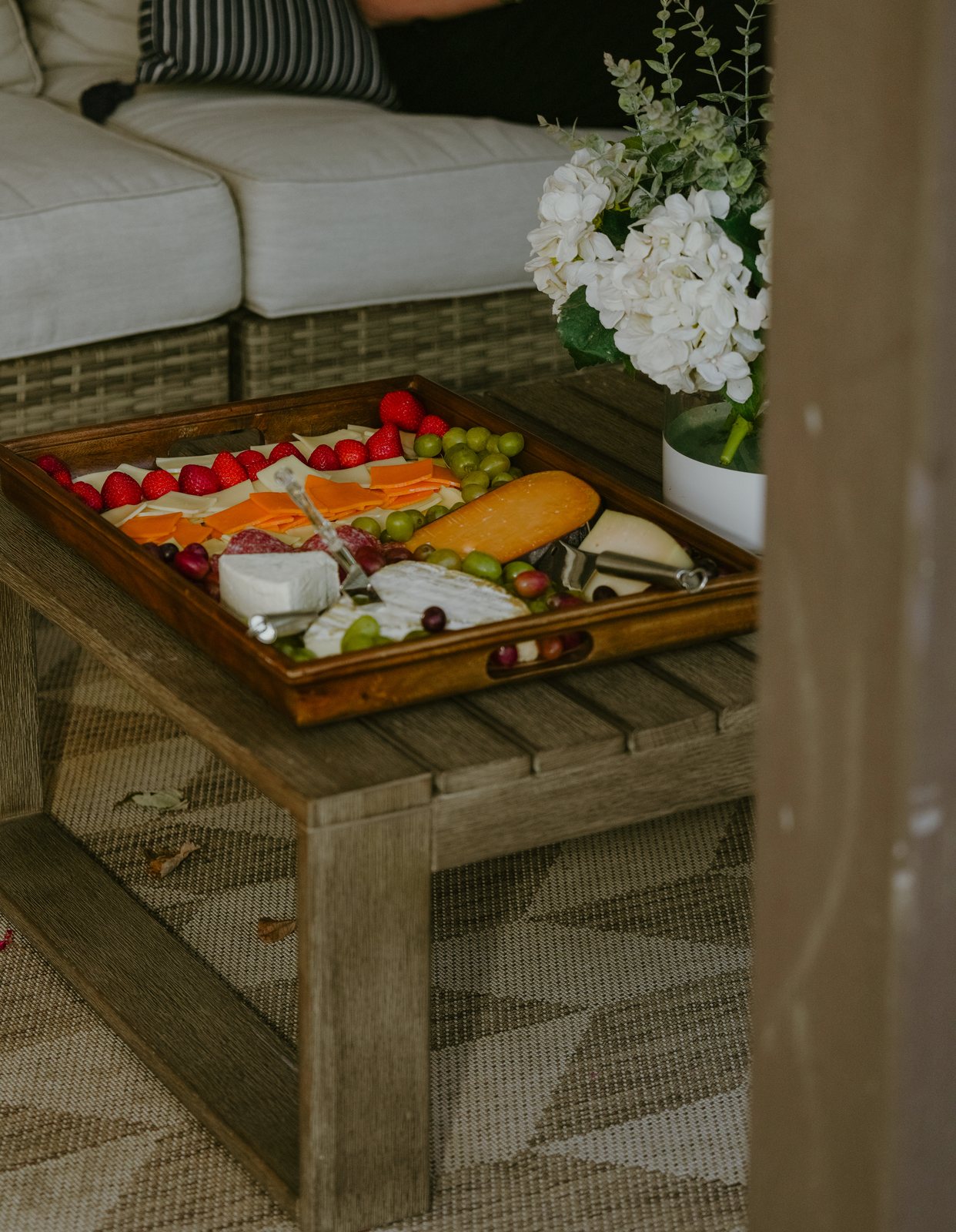 A wooden tray of cheeses, strawberries, grapes and salami sits on a slatted coffee table on the patio, beside white hydrangeas and an outdoor sofa.
