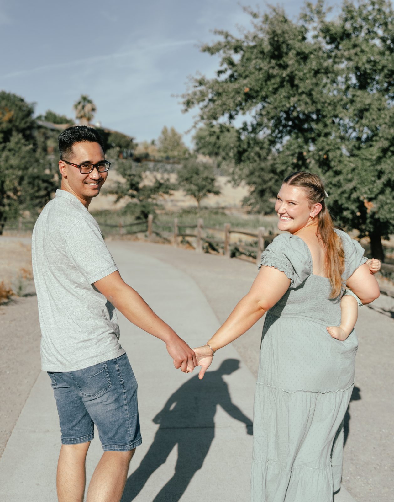 The parents walk away down a paved path holding hands and look back over their shoulders smiling, with the baby on the mother's far side showing only a small hand and a bare foot.