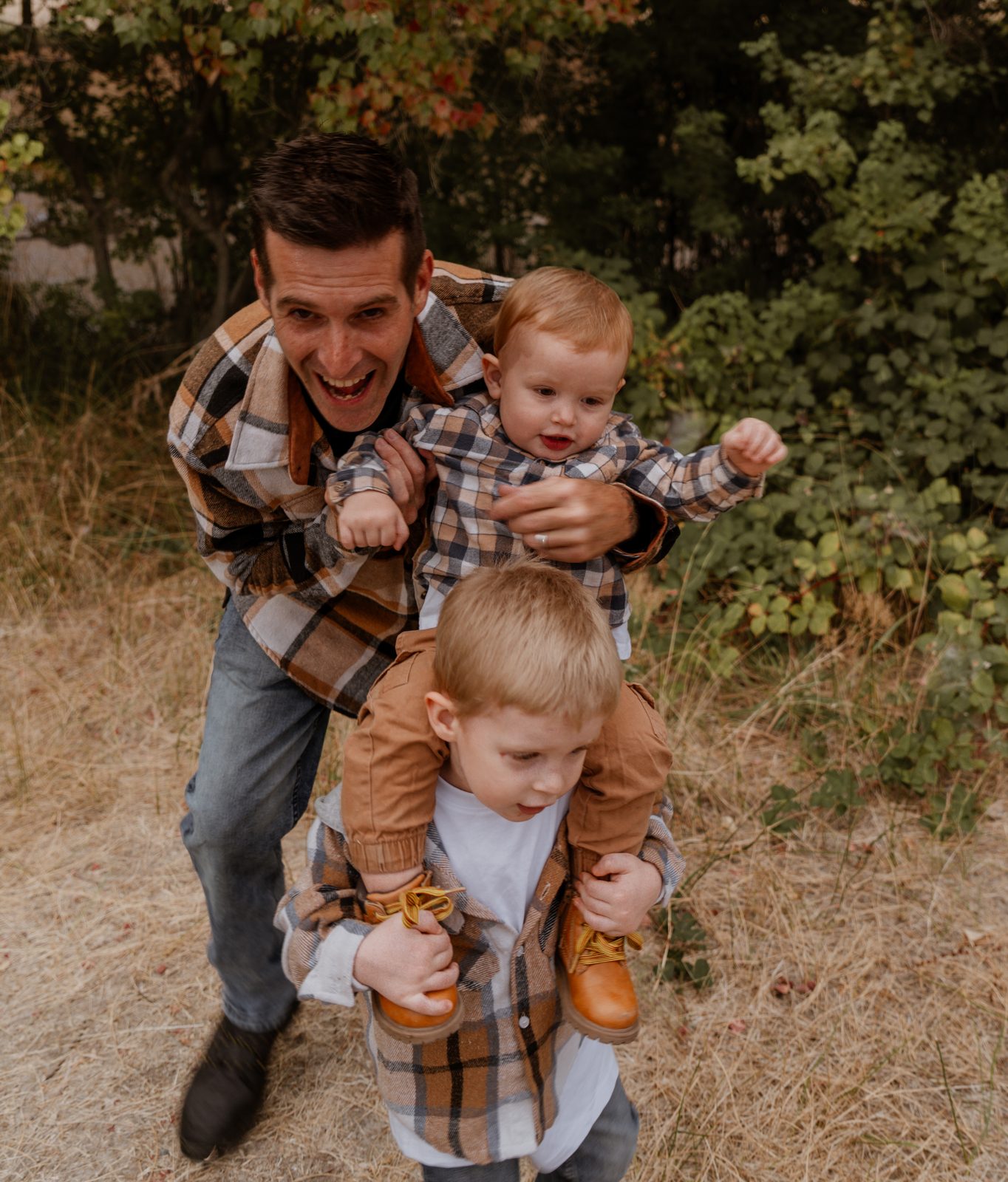 The father, laughing, holds the toddler from behind while the toddler's legs hang over the older boy's shoulders and the older boy grips the toddler's boots.