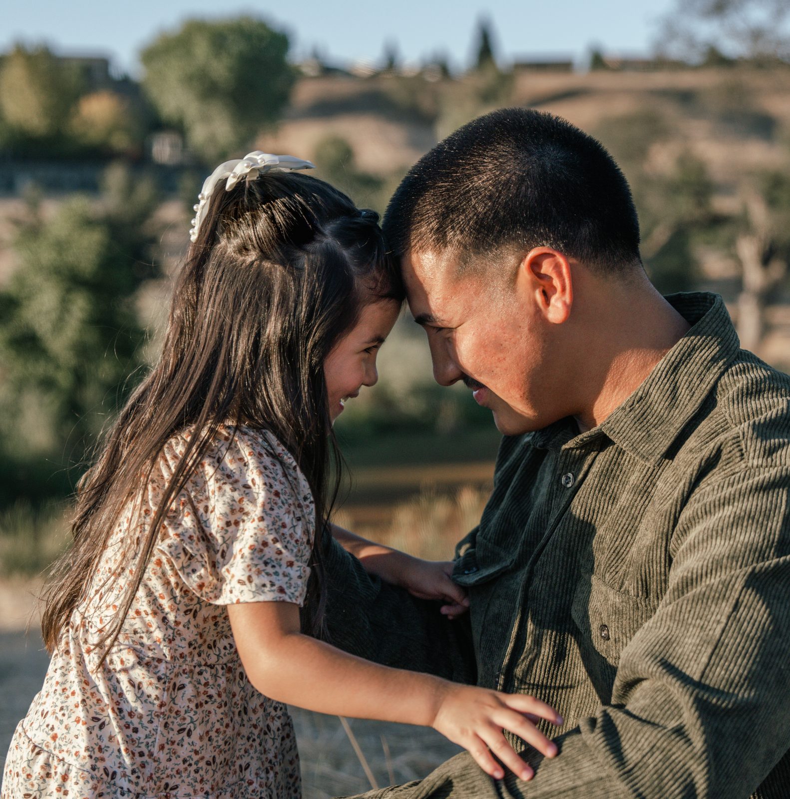 The father and daughter touch foreheads, both smiling with their eyes nearly closed, her hand gripping the front of his green corduroy shirt.