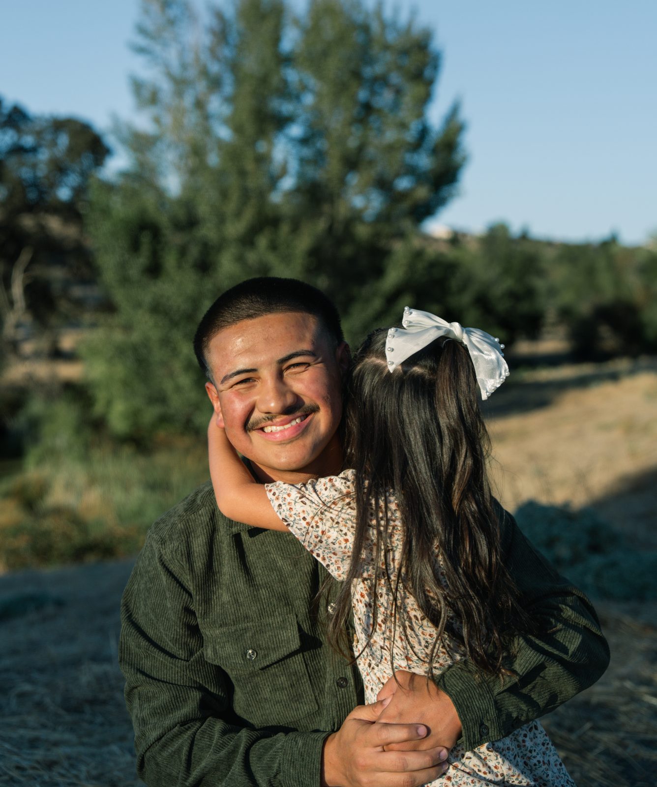 A father grins at the camera as his daughter, a white bow in her hair, wraps her arms around his neck from behind