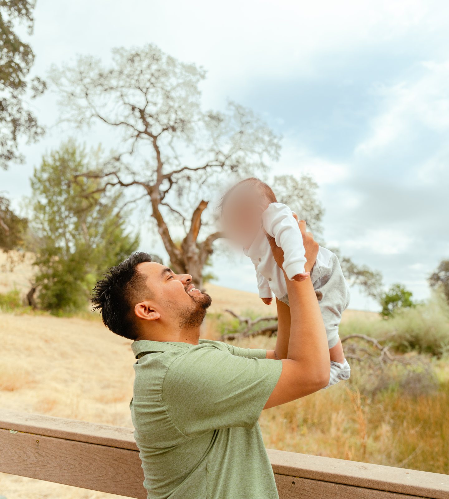 The father grins up at the baby he holds overhead beside a wooden fence rail, with a large oak and dry golden grass behind.