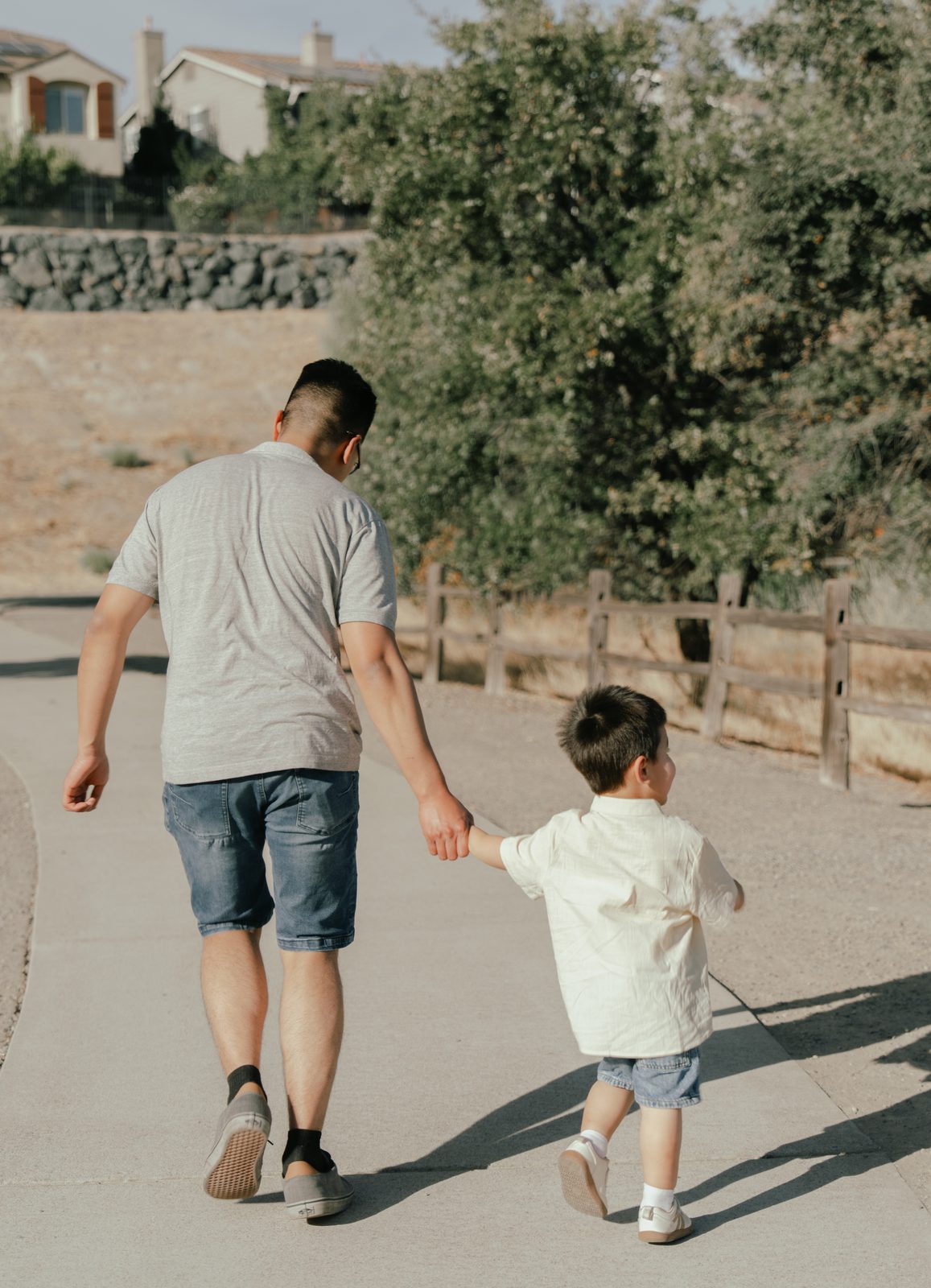 The father and the toddler walk away down a paved path holding hands, beside a wooden fence, with long shadows stretched across the path.