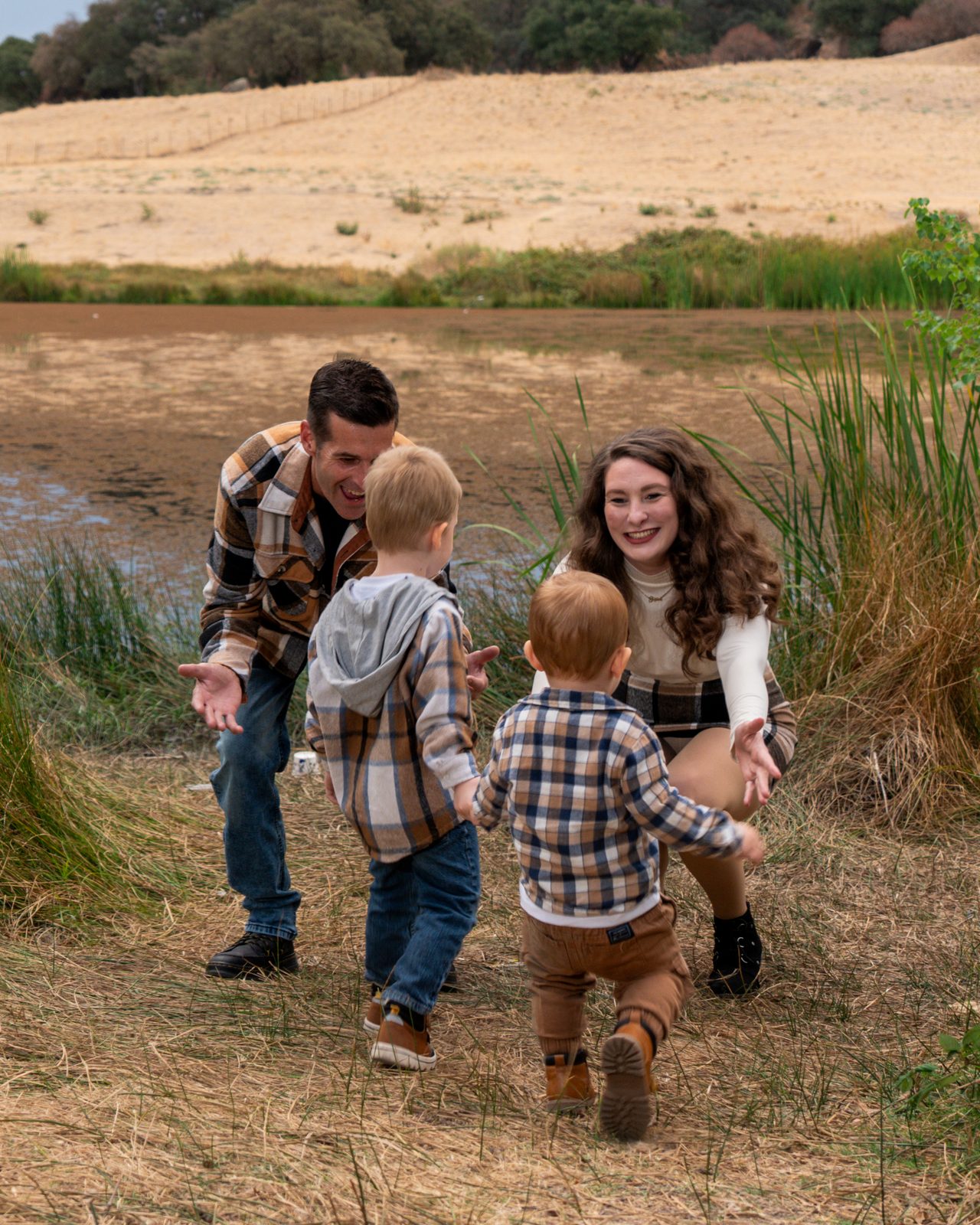 Two small boys in matching plaid toddle across dry grass toward their parents’ open arms