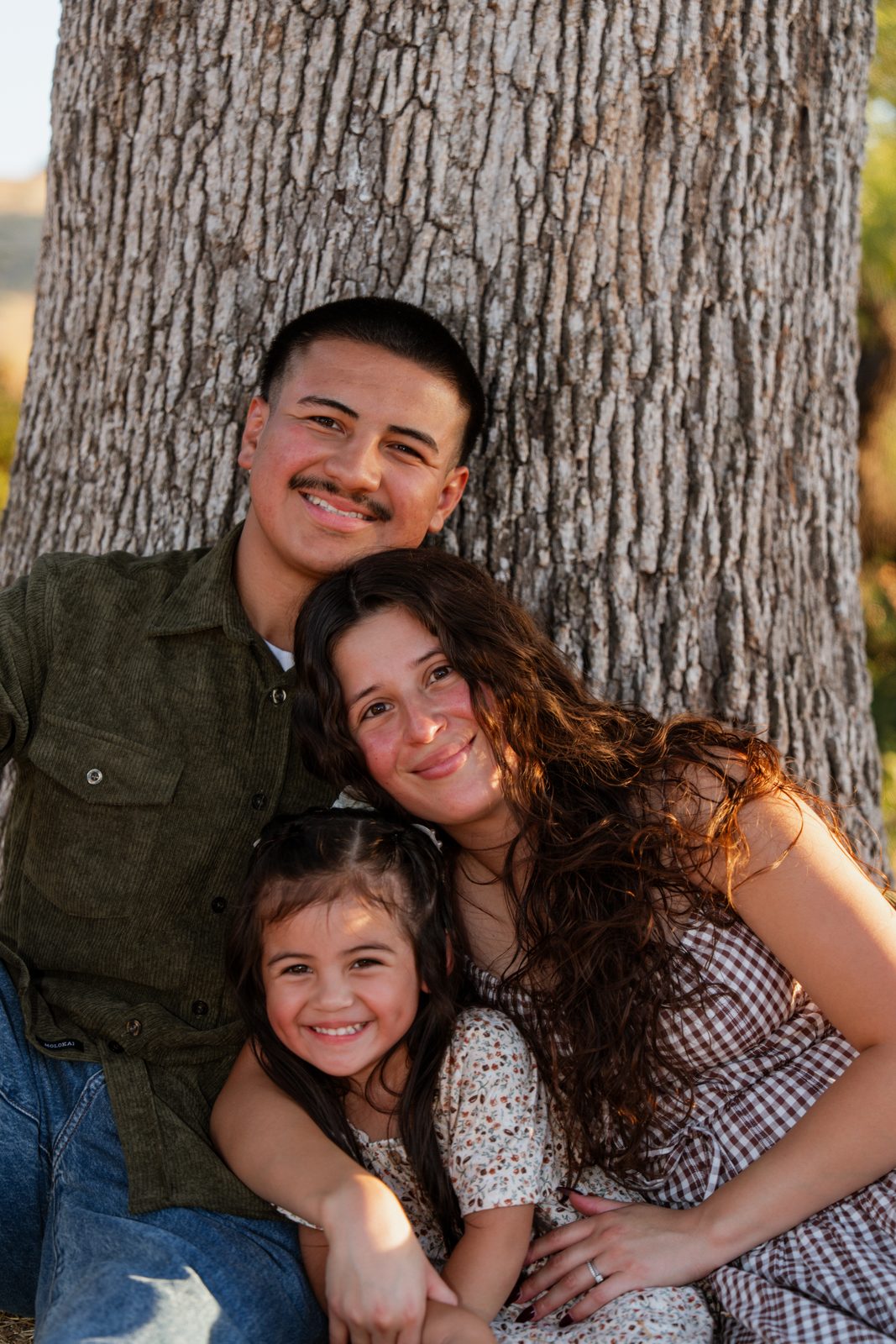 The father, mother and daughter sit pressed close against the trunk of an oak, their heads stacked one above the other, all three smiling at the camera.