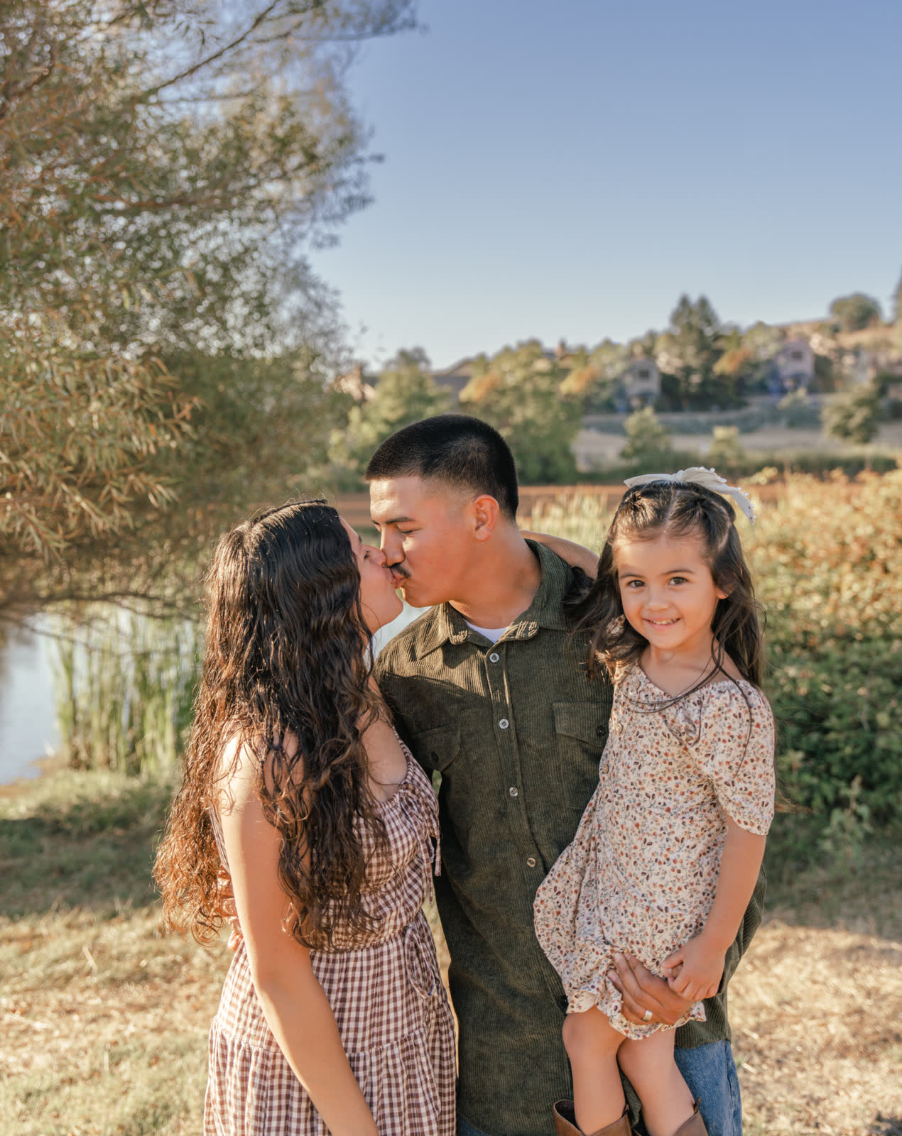 The father, holding his daughter on his hip, kisses the mother while the girl grins at the camera, beside a reedy pond with houses on the hill behind.