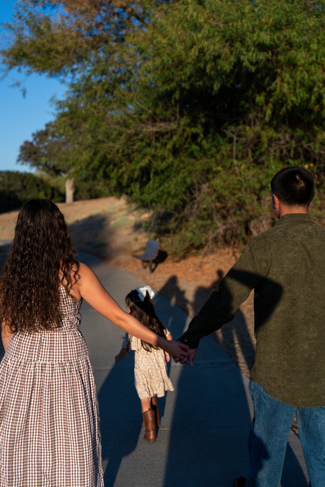 Seen from behind, the mother and father hold hands while their daughter in a floral dress and cowboy boots walks ahead of them down a paved path crossed by long shadows, toward a bench under a tree.
