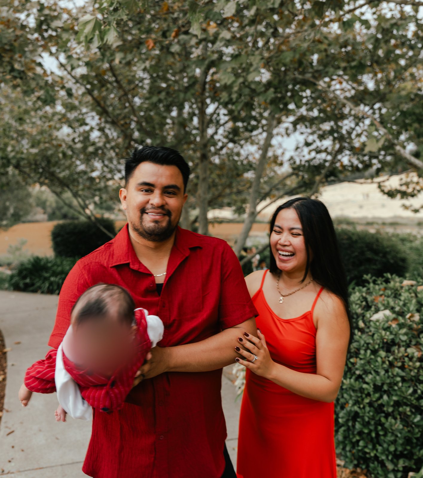 The father smiles at the camera holding the baby out along his arm in a red striped outfit, while the mother laughs and holds onto his arm on a tree-lined path.