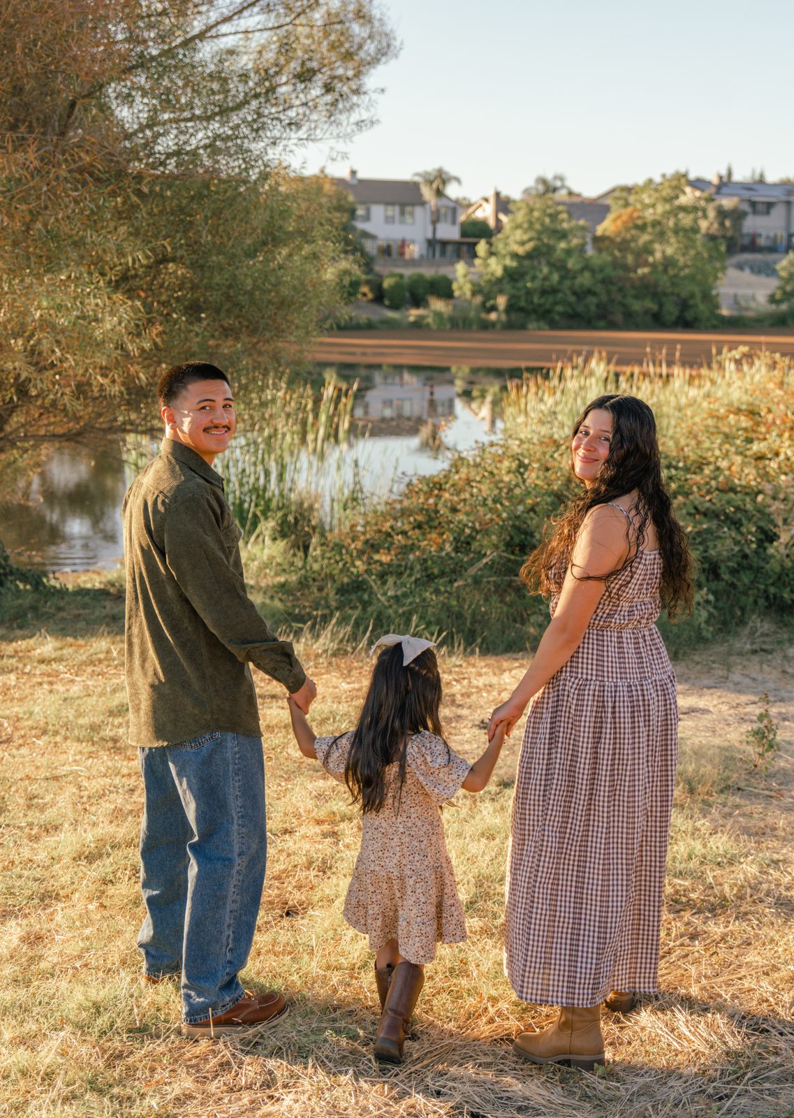 With their backs to the camera, the parents hold their daughter's hands between them in dry grass facing a reed-lined pond, both looking back over their shoulders and smiling.