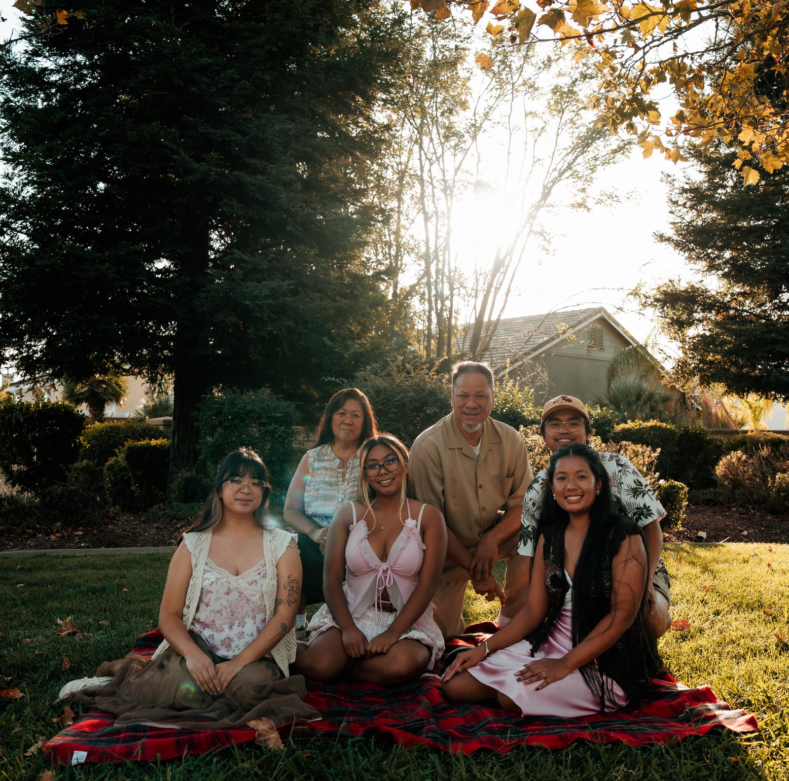 Six family members sit together on a red plaid blanket on the grass, the parents in the back row, with tall evergreens, a house roof and the sun shining through the trees behind them.