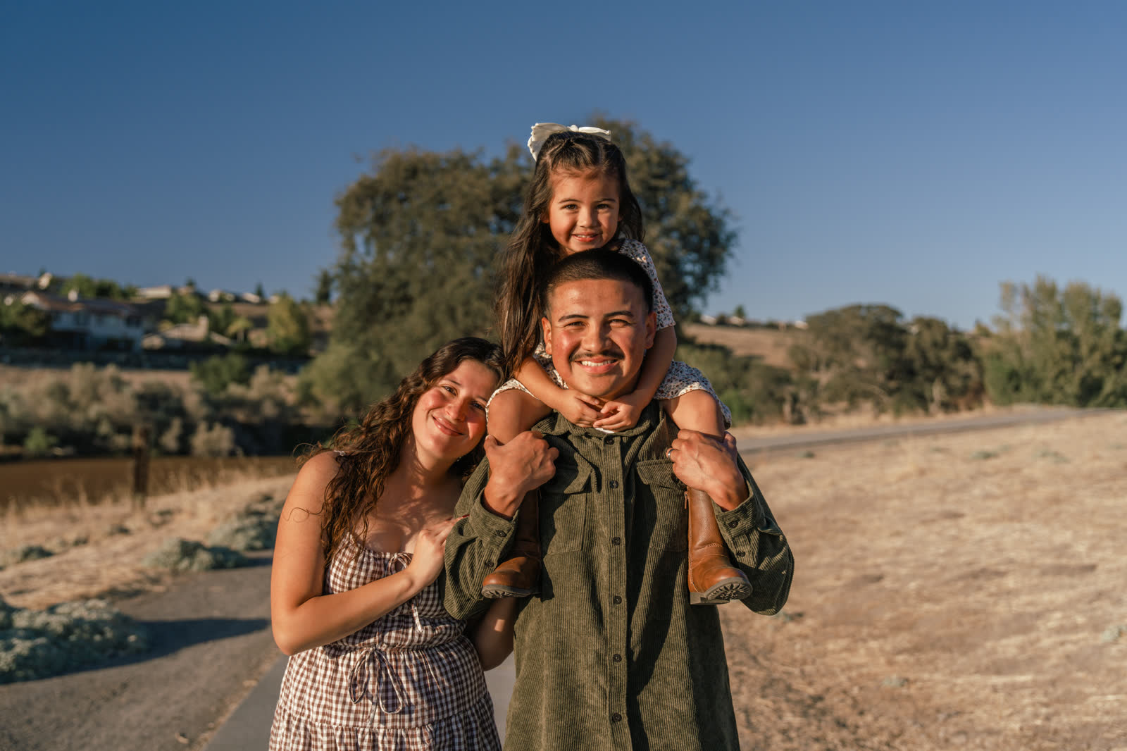 The father grins at the camera with his daughter on his shoulders, her hands clasped under his chin and his hands holding her legs, while the mother leans in beside him on a path through dry grass with an oak and blue sky behind.