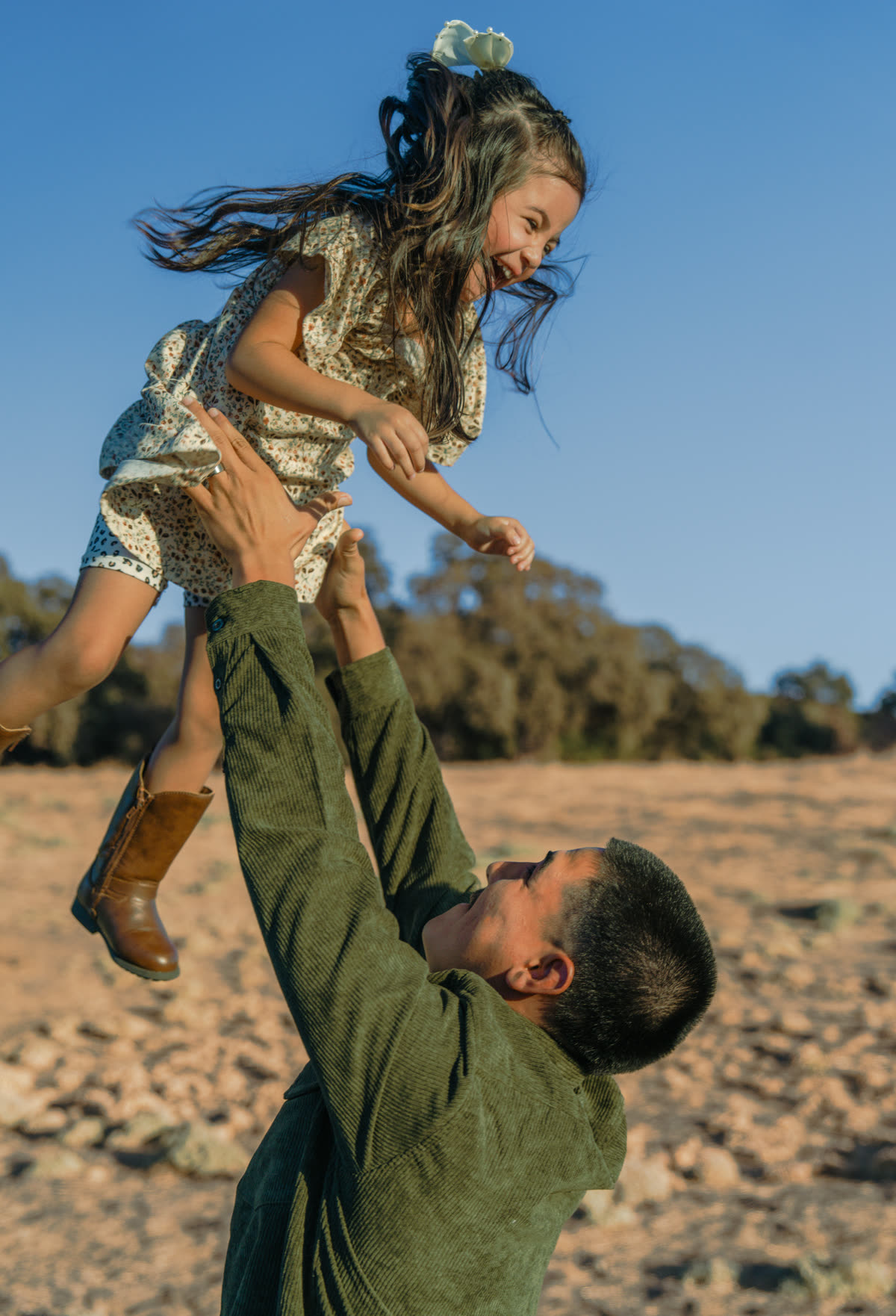 The daughter up in the air above her father's head, laughing with her hair flying out, as he reaches up to her with both hands in a sunlit field below a line of oaks.