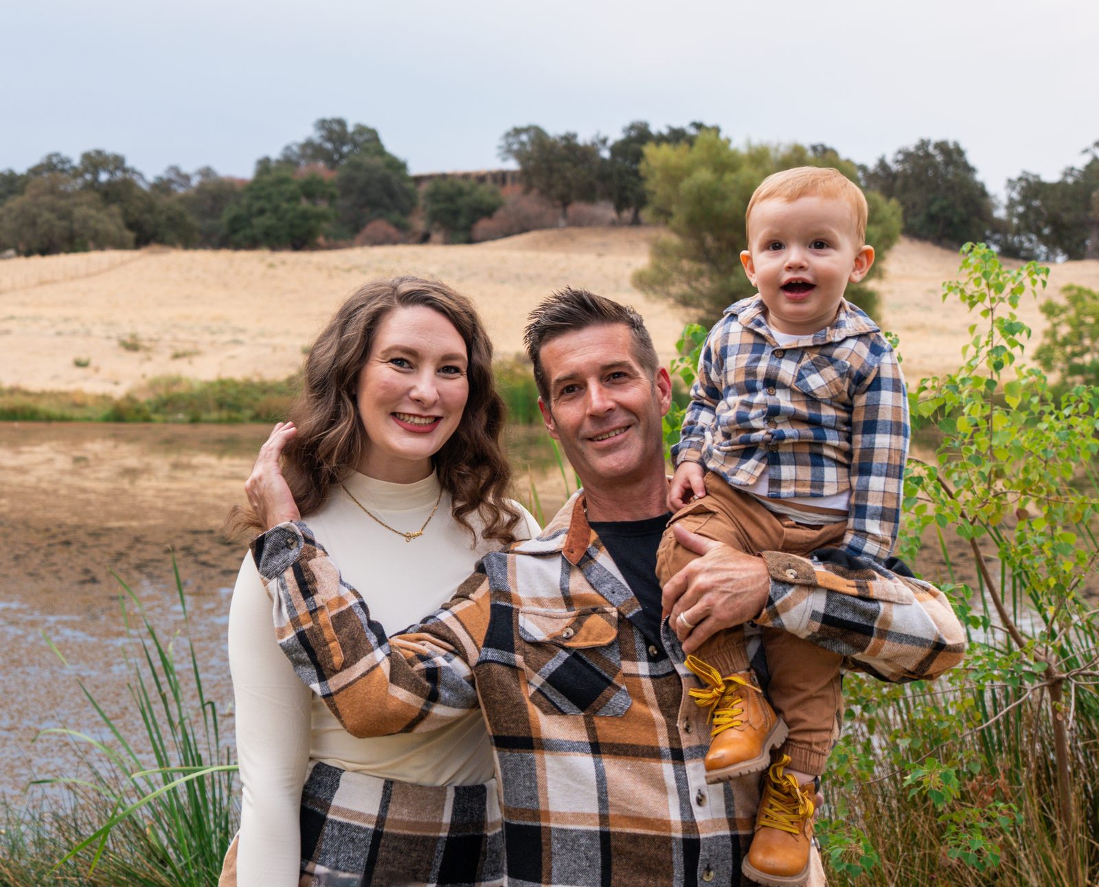 The parents smile at the camera beside the pond while the father holds the grinning toddler up on one arm.